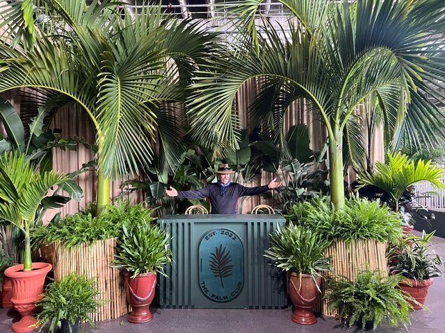 Man stands behind teal bar with palm trees, surrounded by plants; tropical setting.