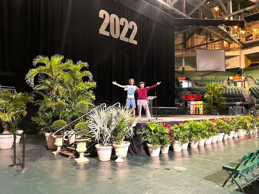 Two people on a decorated stage with "2022" backdrop, surrounded by plants.