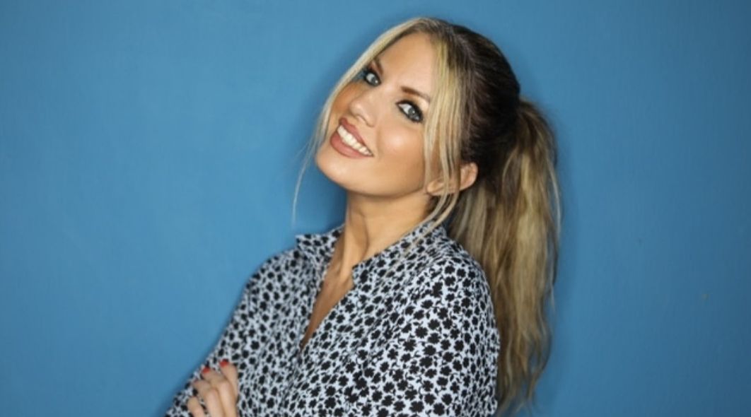 Une femme aux cheveux blonds coiffés en queue de cheval sourit, les bras croisés, vêtue d'une chemise à motifs sur fond bleu.