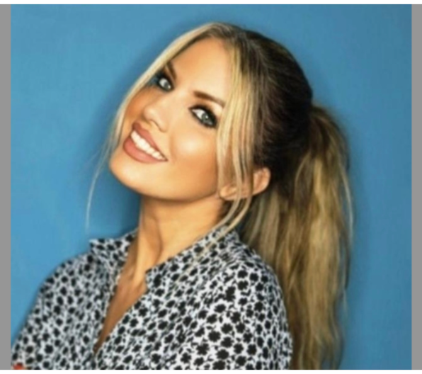 Une femme aux cheveux blonds coiffés en queue de cheval sourit, les bras croisés, vêtue d'une chemise à motifs sur fond bleu.
