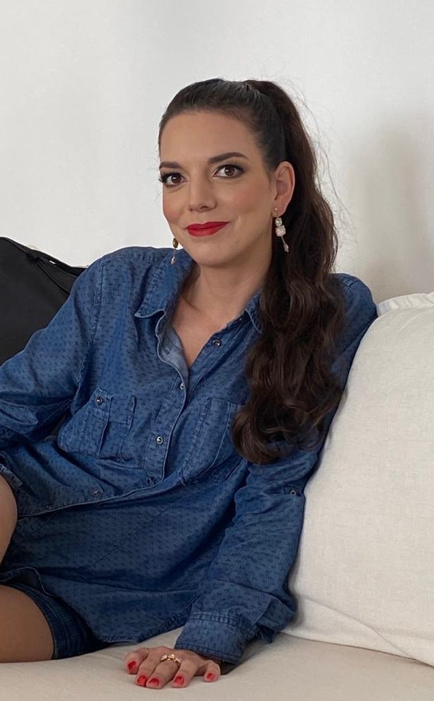 Femme en chemise et short en jean, rouge à lèvres rouge, queue de cheval, assise sur un canapé avec des coussins, souriante.