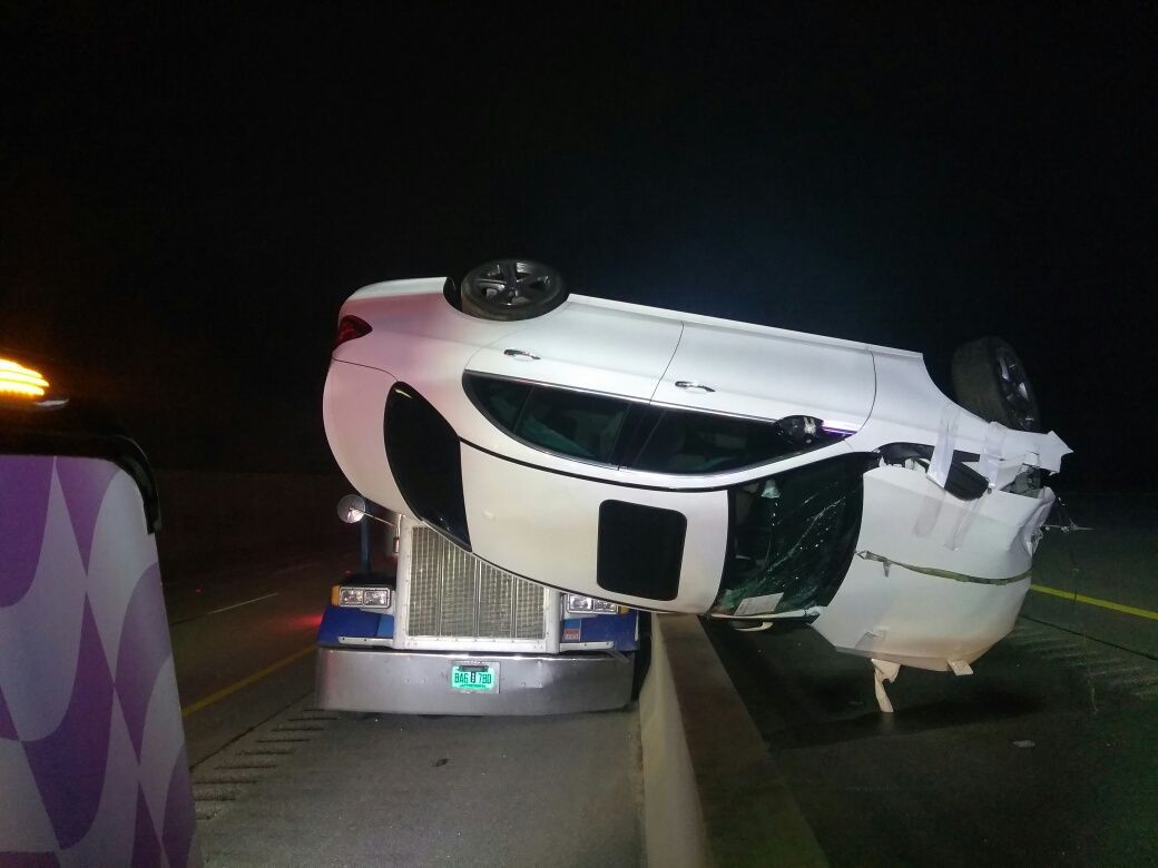 White car flipped over on highway, being towed by a semi-truck at night.