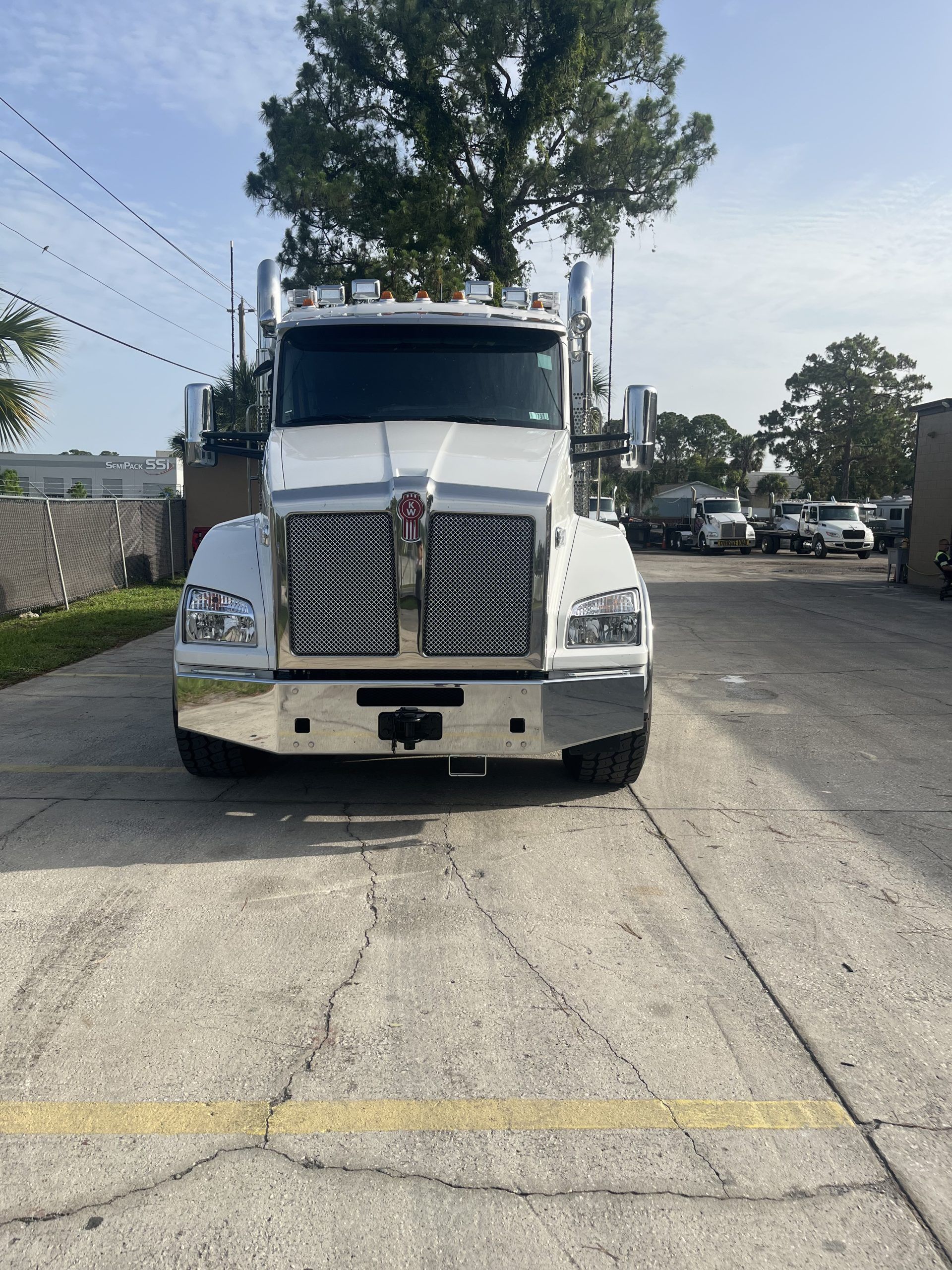 White Kenworth semi-truck parked on concrete, facing camera, with a tree and other vehicles in the background.