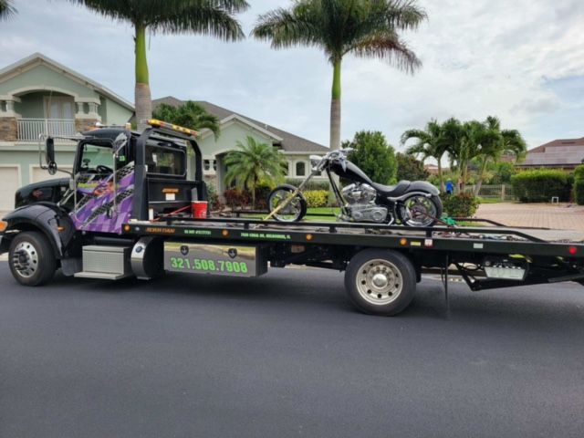 Black tow truck transporting a custom motorcycle on a residential street.