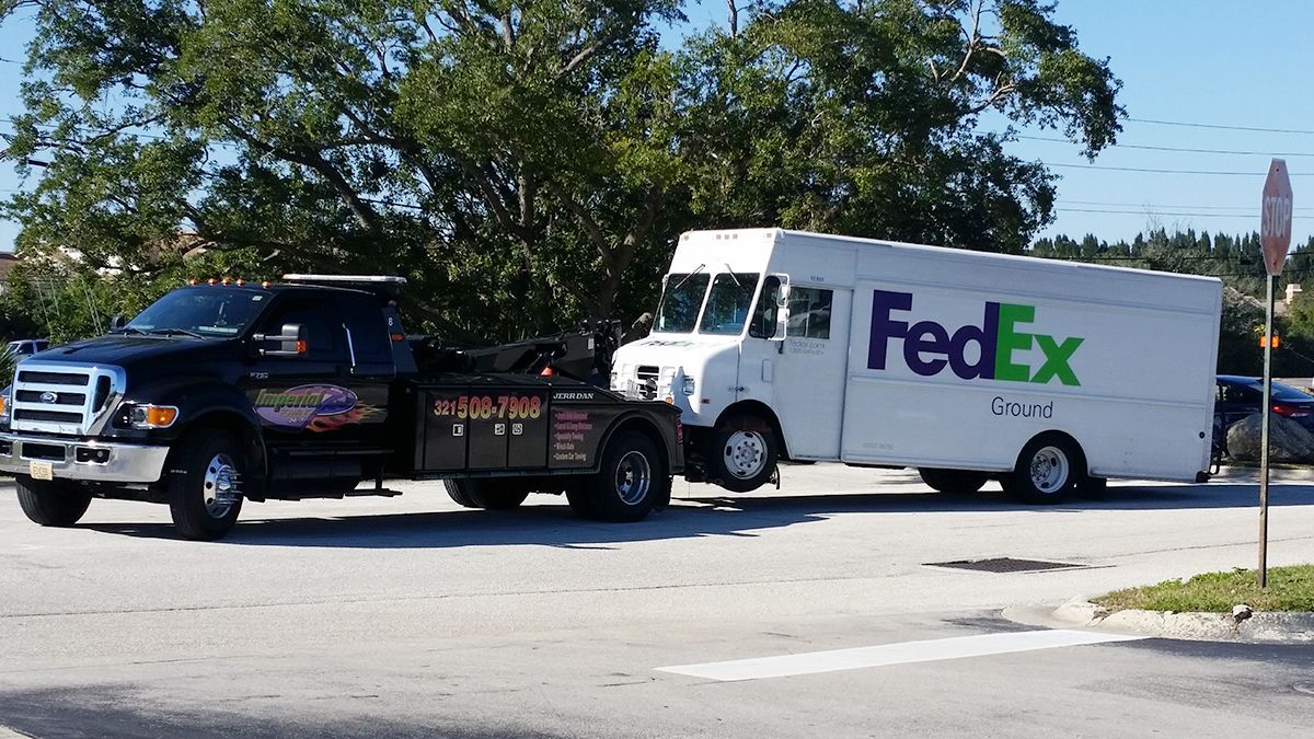 Black tow truck towing a white FedEx delivery truck on a road; the vehicles are side-by-side.