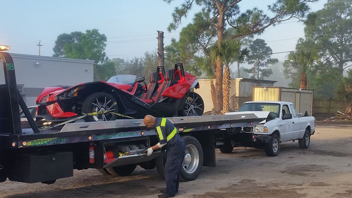 A damaged red Polaris Slingshot on a tow truck. A worker adjusts the truck, a white pickup is also attached.