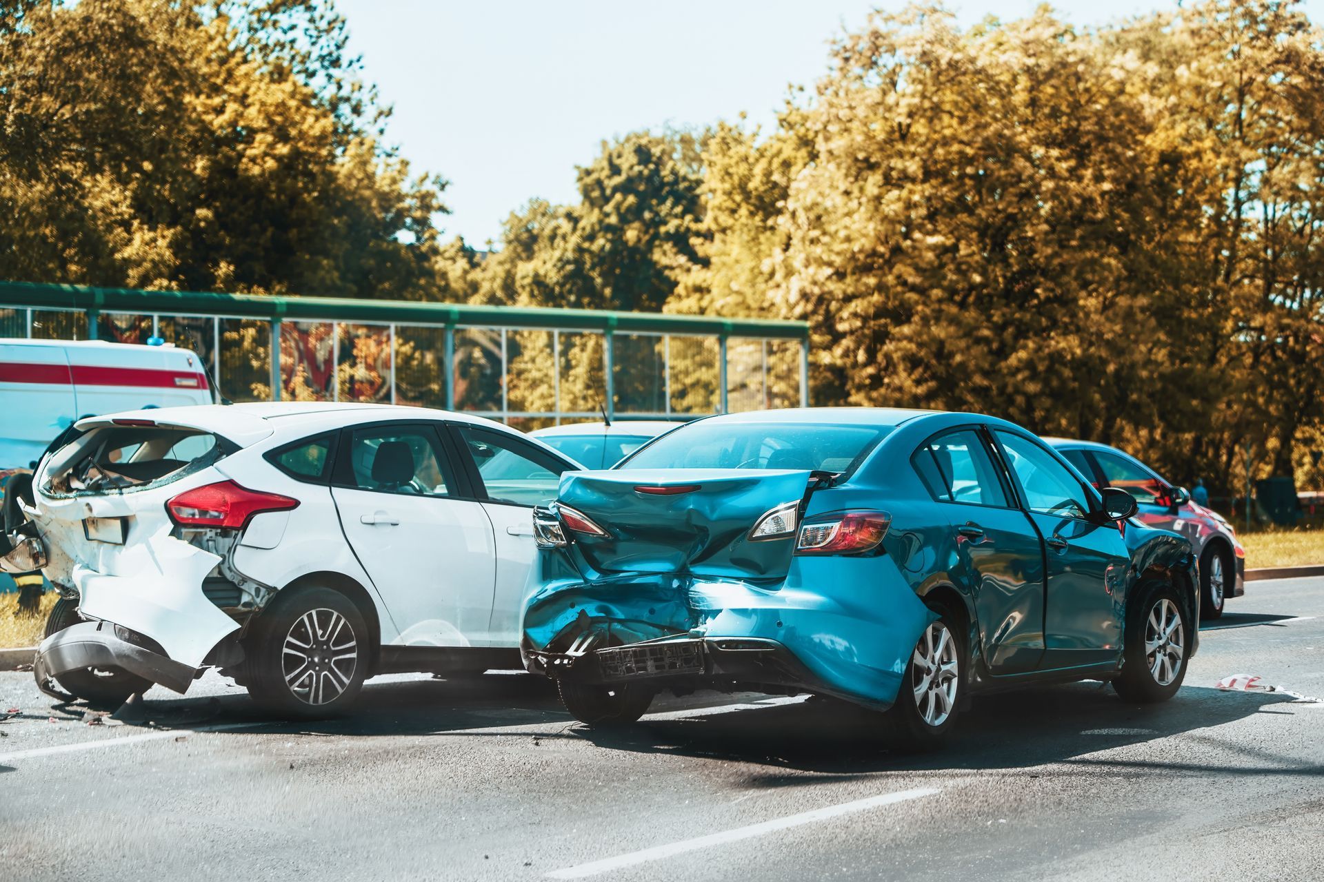 Two cars with severe rear-end collision damage on a road, with trees and a guardrail in the background.