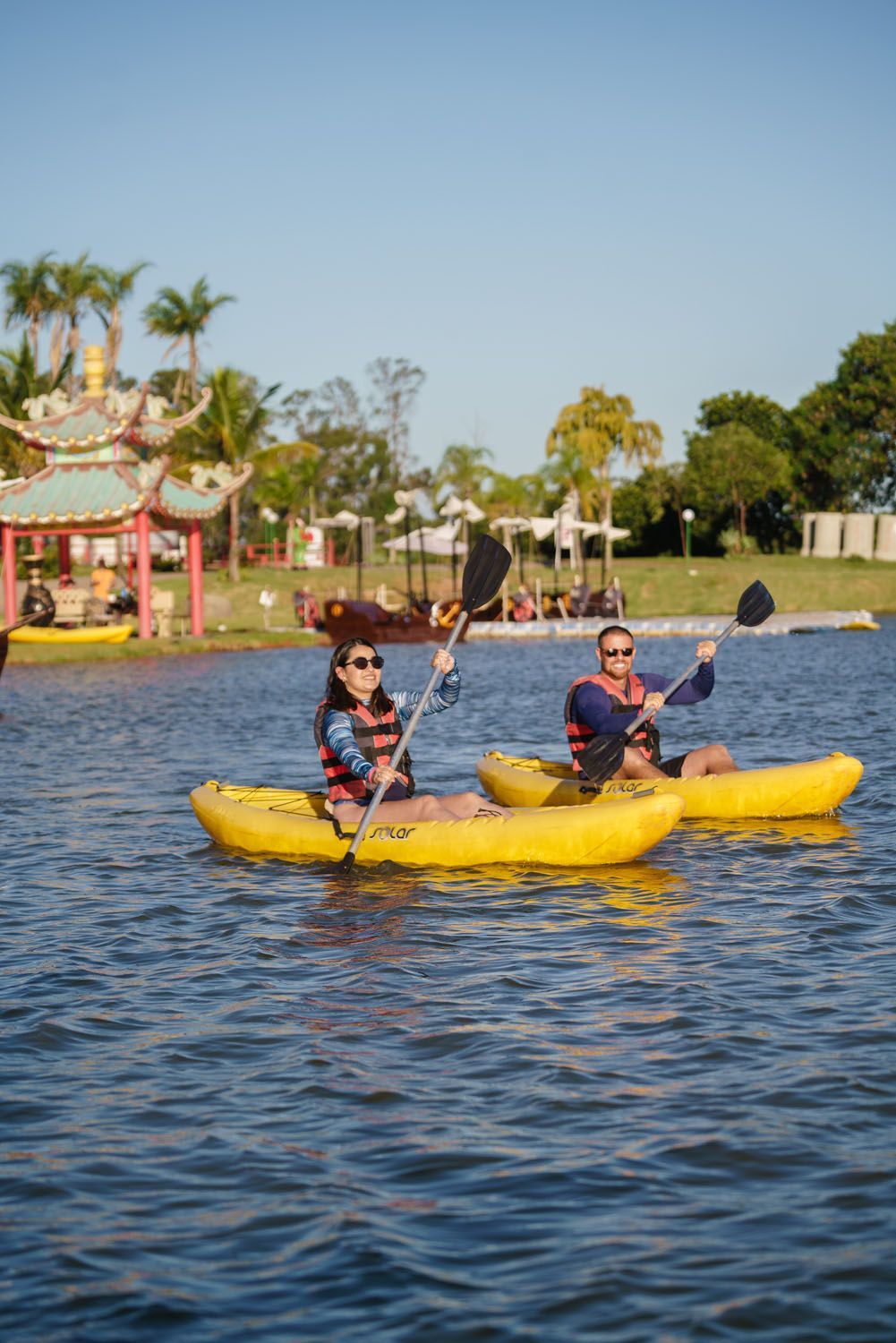 Duas pessoas andam de caiaque em um lago perto de uma construção em estilo pagode. Elas usam coletes salva-vidas e remam.