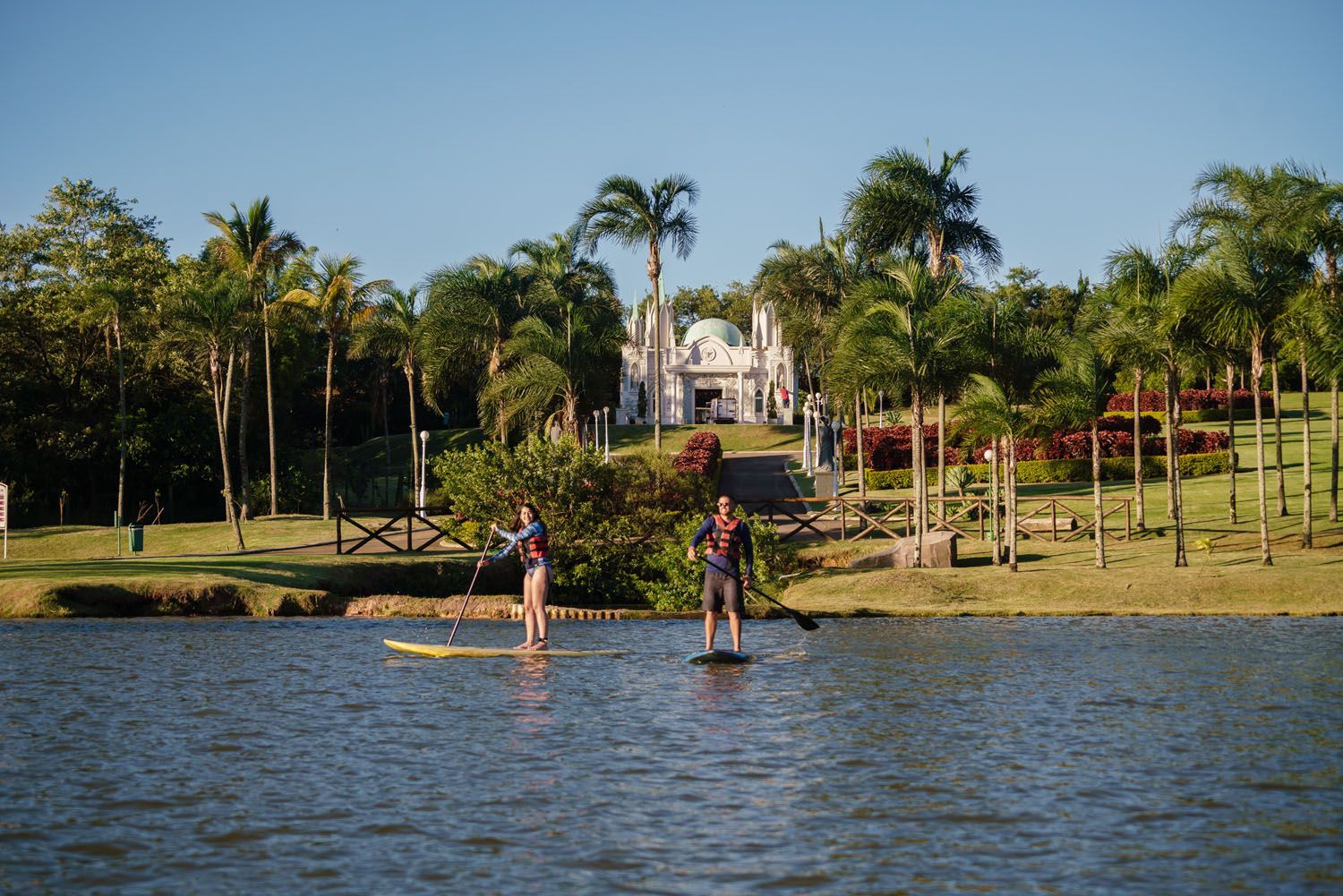 Duas pessoas praticam stand up paddle em um lago, com árvores e um prédio branco ao fundo.