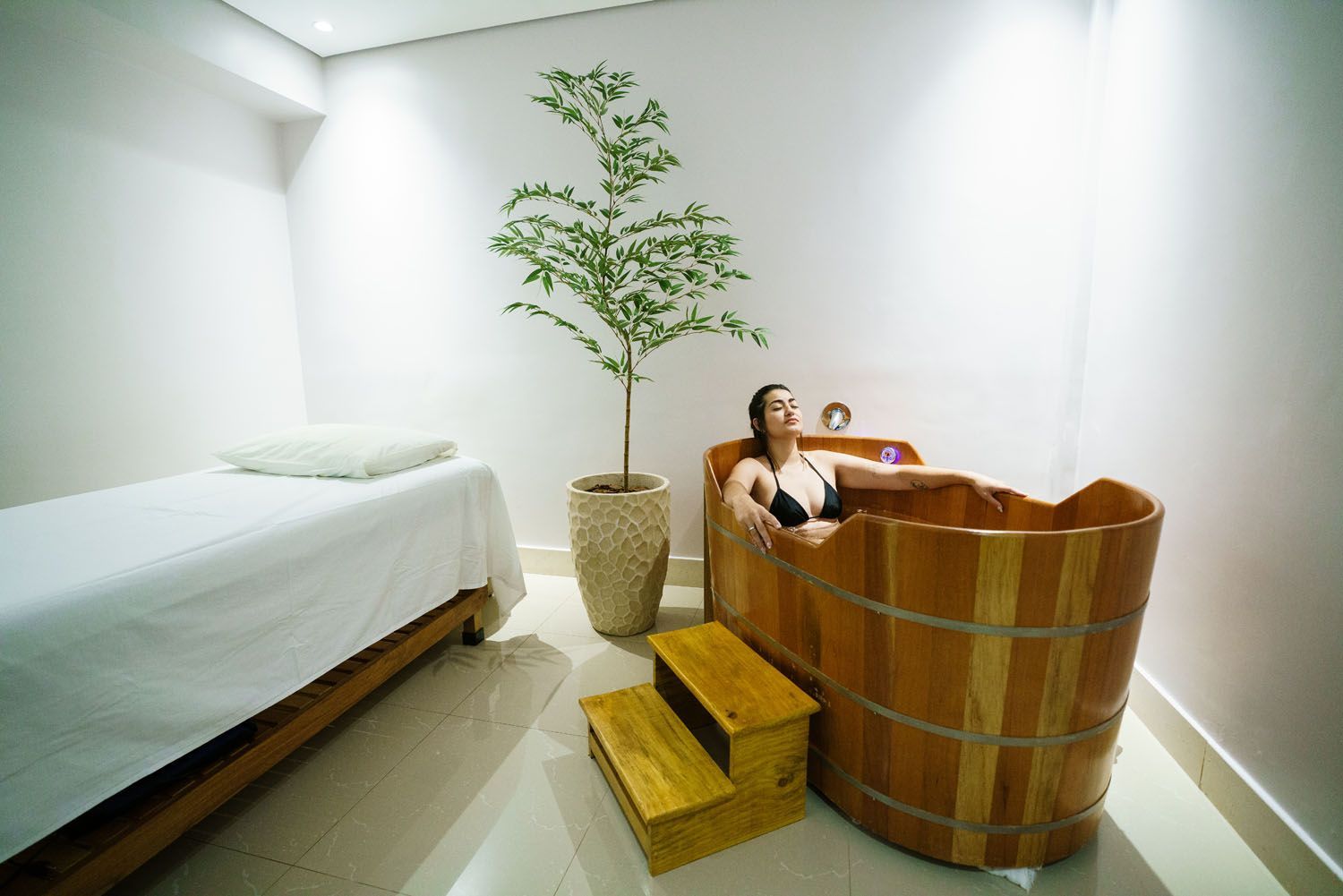 Mulher em banheira de madeira, ambiente de spa com cama de massagem, planta e degraus de madeira.