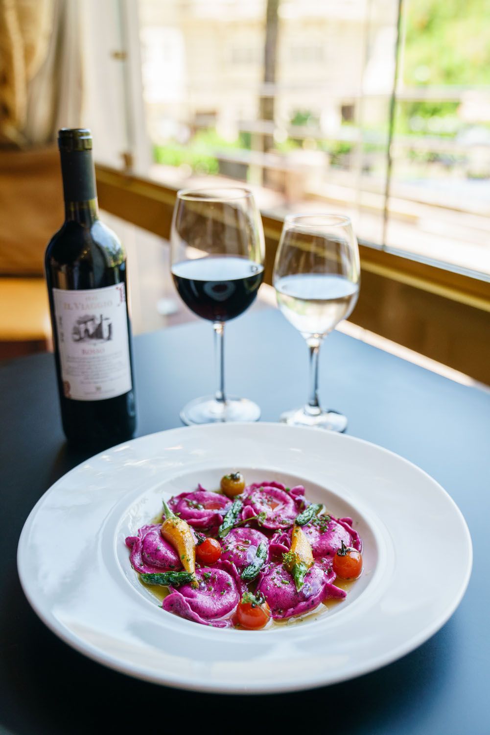 Prato de ravióli roxo, duas taças de vinho e uma garrafa de vinho tinto perto de uma janela.