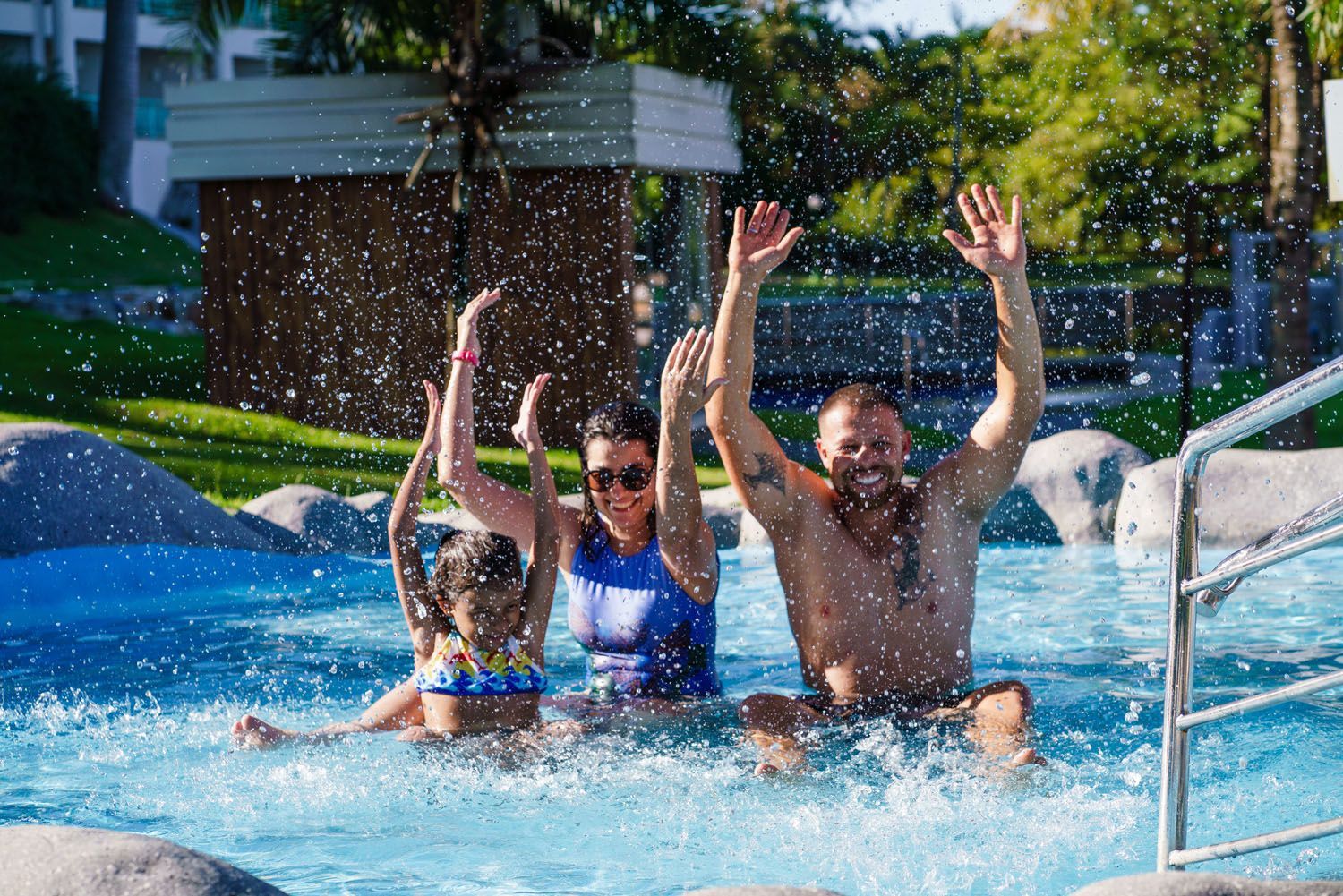 Família brincando na piscina, braços erguidos, empolgados. Luz do sol, água azul e grama verde ao fundo.