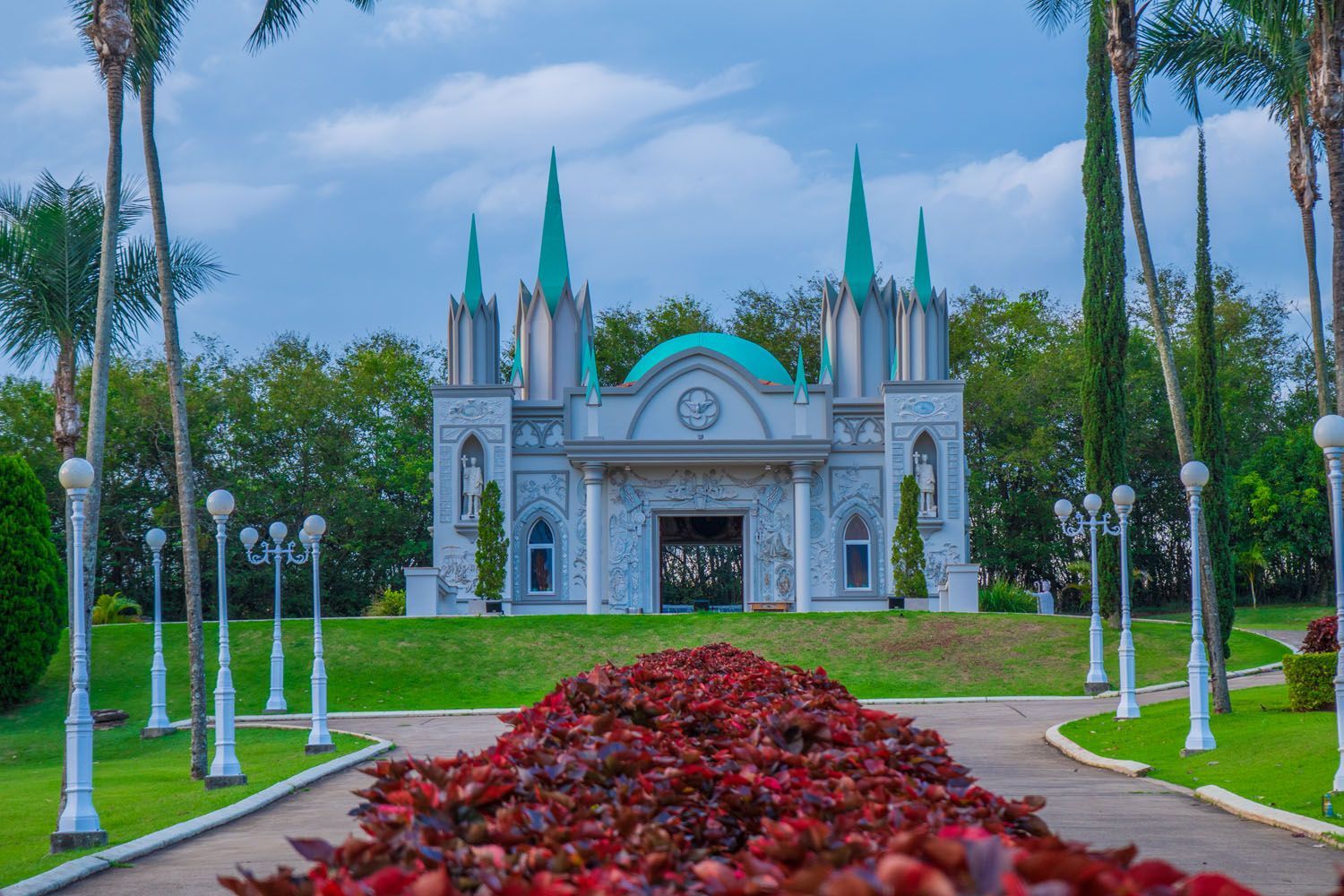 Mausoléu com torres e cúpula azul-petróleo, com folhagem vermelha na frente e ladeado por árvores, sob um céu nublado.