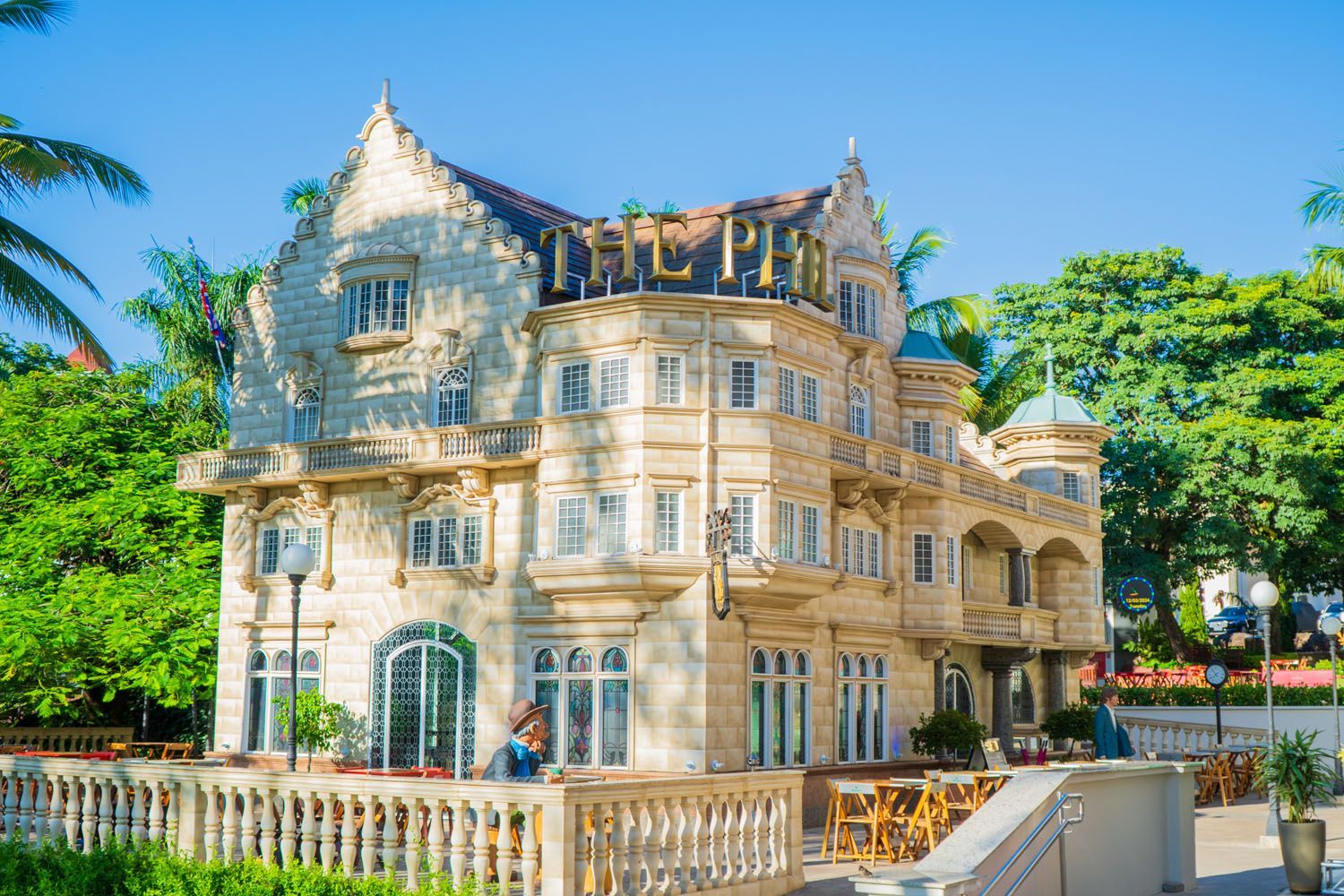 Mansão bege de vários andares com detalhes ornamentados, cercada por árvores e uma balaustrada em um dia ensolarado.