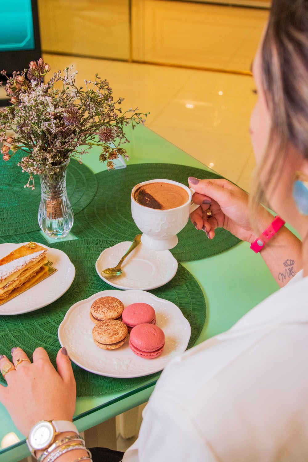 Mulher segurando uma xícara de café; mesa com macarons, churros e flores.