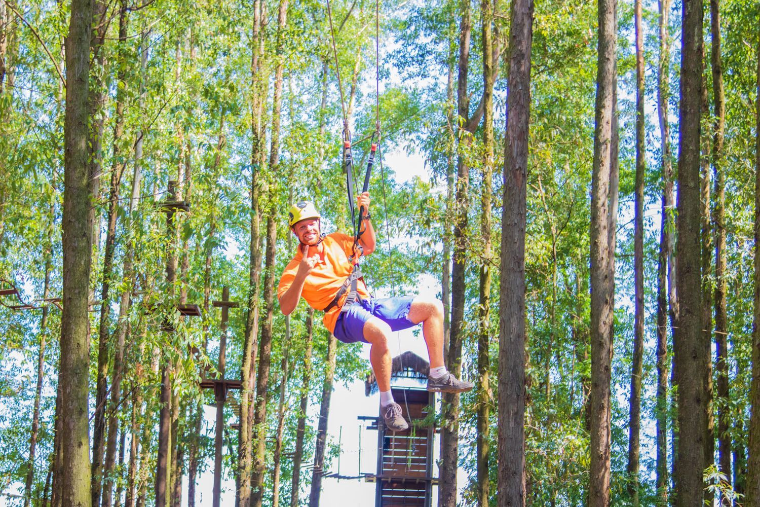 Homem em uma tirolesa, vestindo camisa laranja e shorts azuis, em uma floresta.