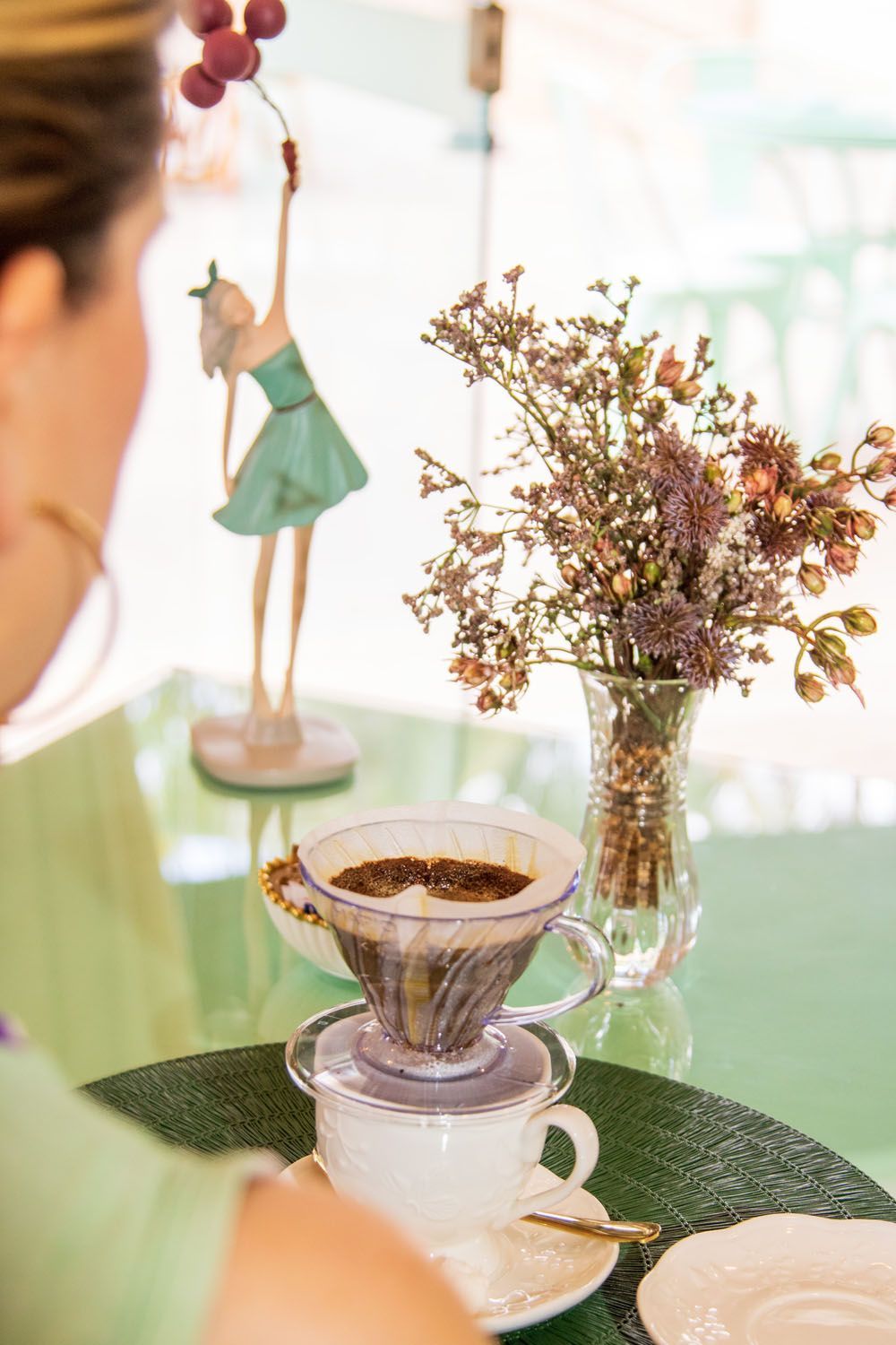 Mulher perto de um recipiente de café, flores e uma estatueta em uma mesa verde em uma sala iluminada.