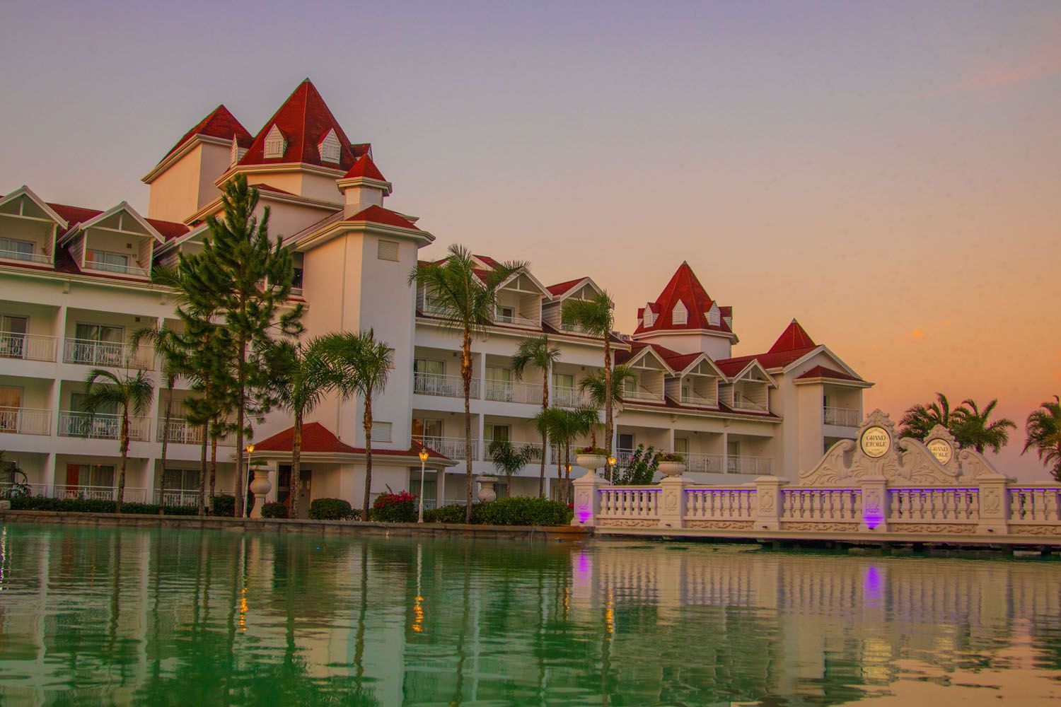 Edifício branco do resort com torres de telhado vermelho refletido em uma piscina verdejante ao pôr do sol.