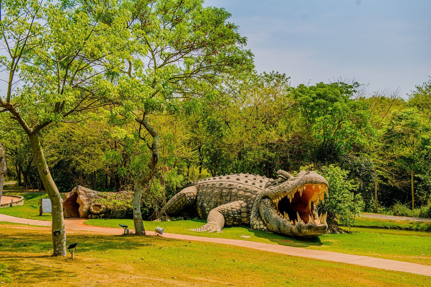 Estátua gigante de crocodilo com a boca aberta em um gramado cercado por árvores.
