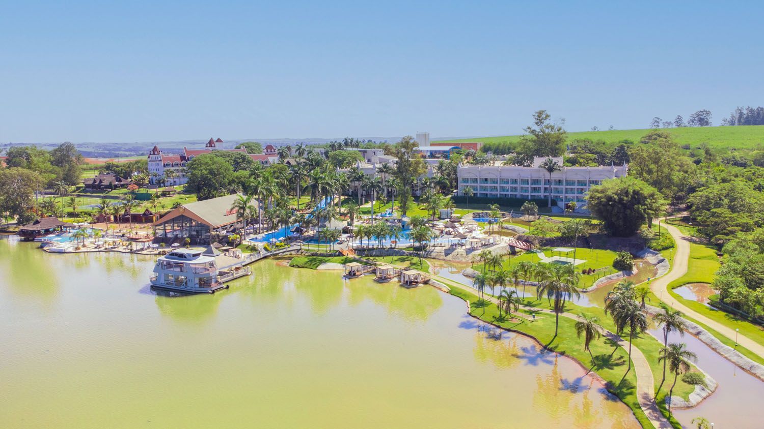 Complexo de resort à beira de um lago, com prédios, piscinas, palmeiras e paisagens verdes sob um céu azul claro.