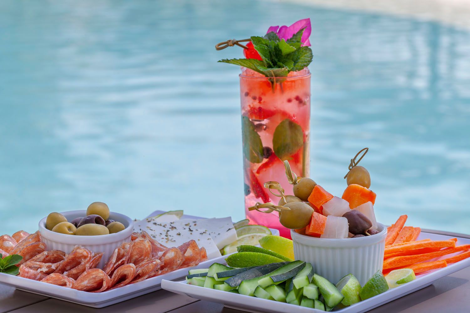 Mesa de coquetéis e frios à beira da piscina. Inclui azeitonas, carnes, legumes e espetinhos.