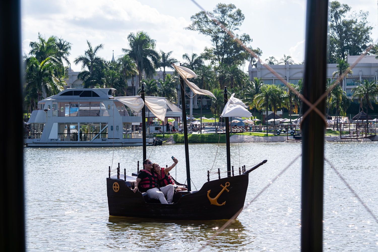 Casal em um pequeno navio pirata na água, tirando uma selfie. Barco branco, palmeiras ao fundo.