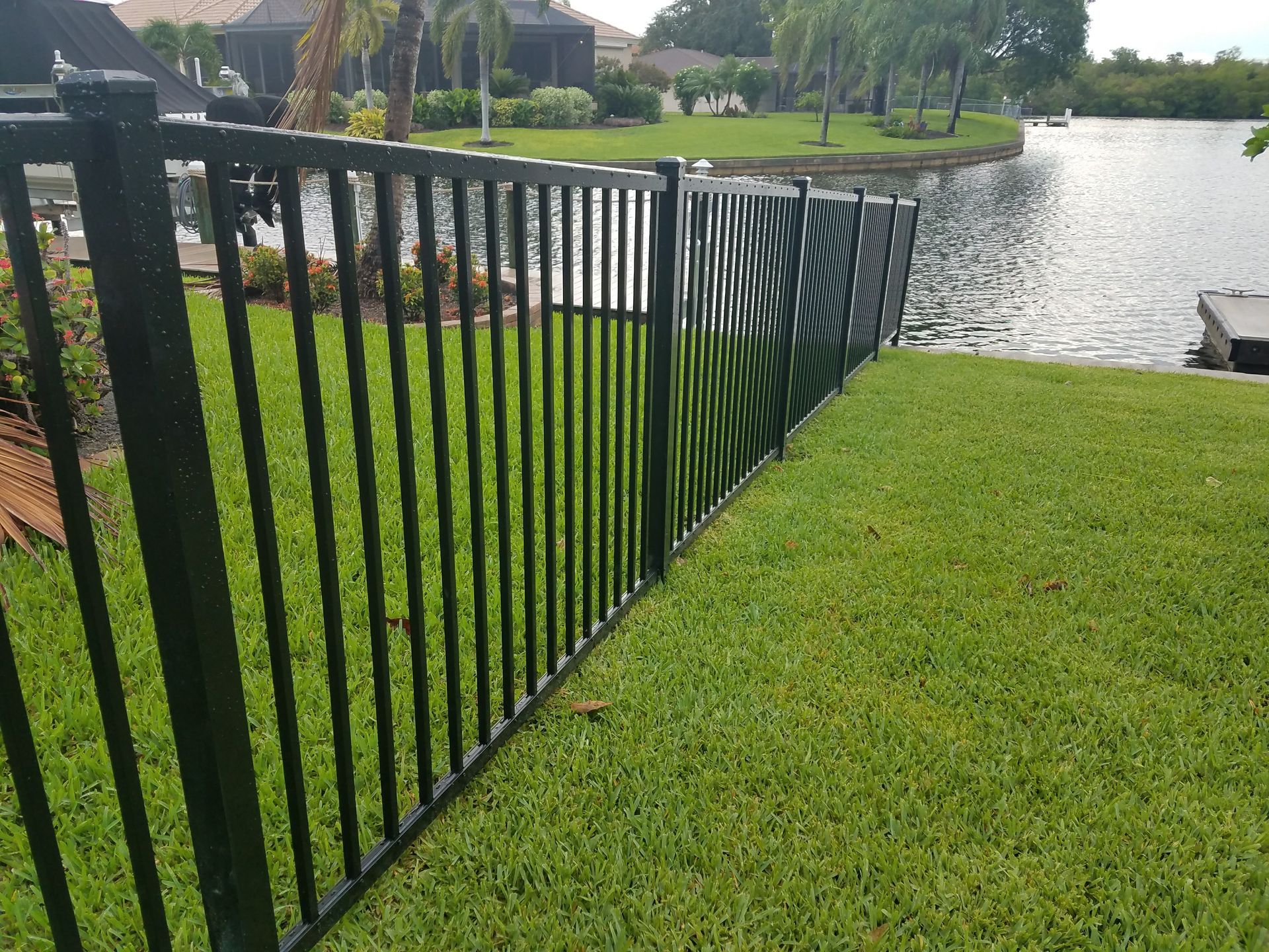 A black fence surrounds a lush green lawn next to a body of water.