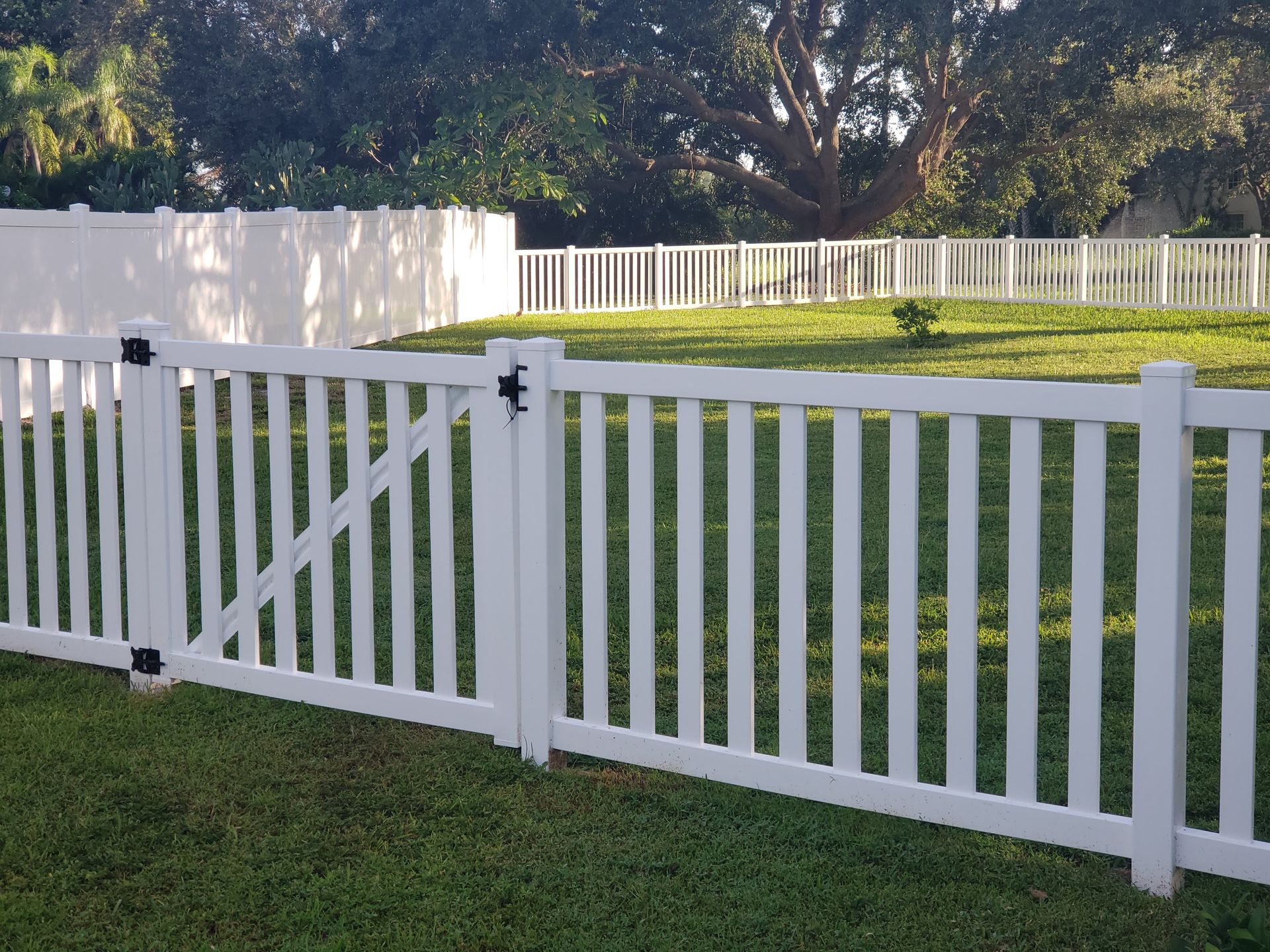 White picket fence with gate in a grassy yard, trees in the background.