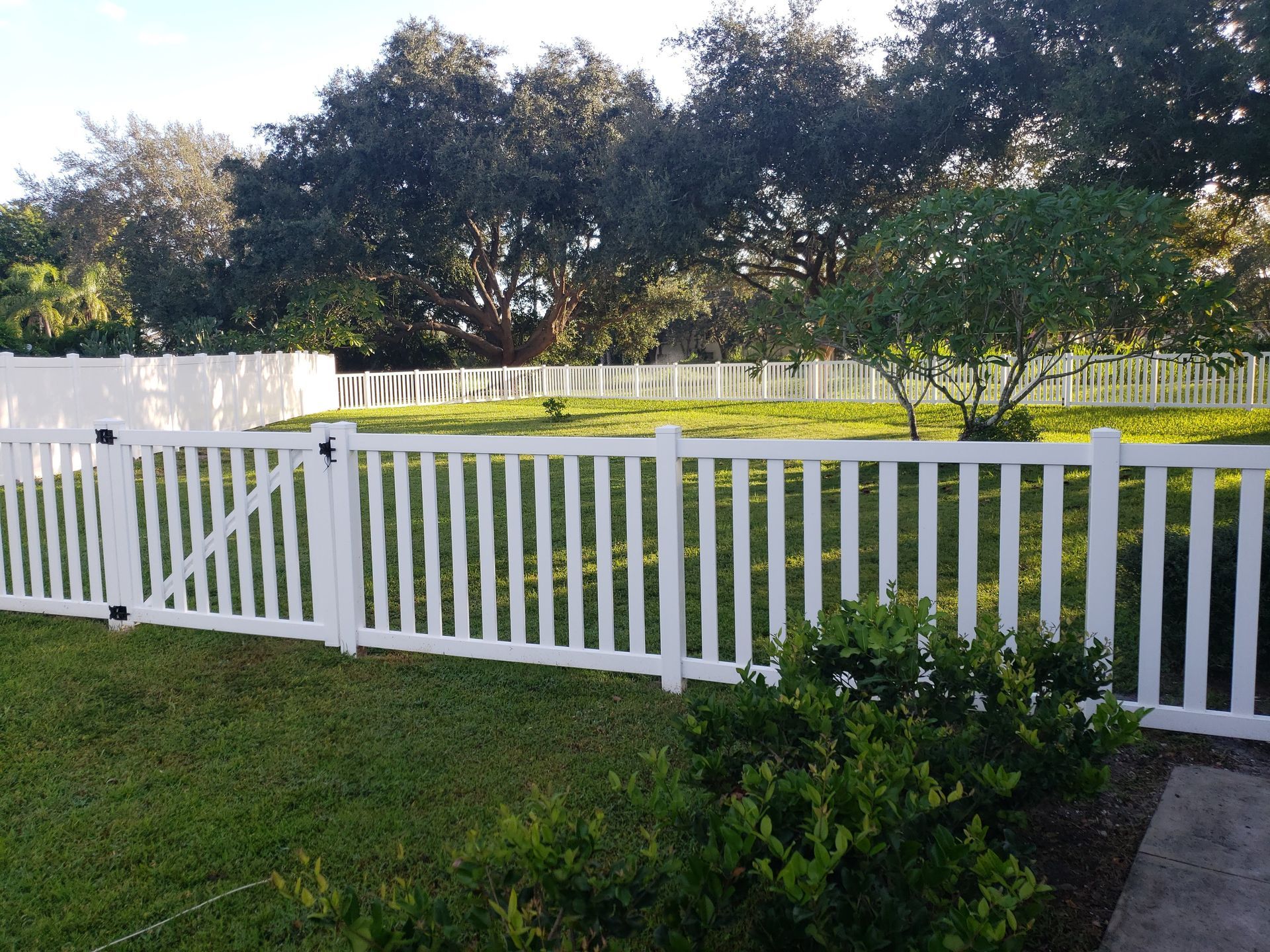 White picket fence encloses a green lawn with trees in the background under a sunny sky.