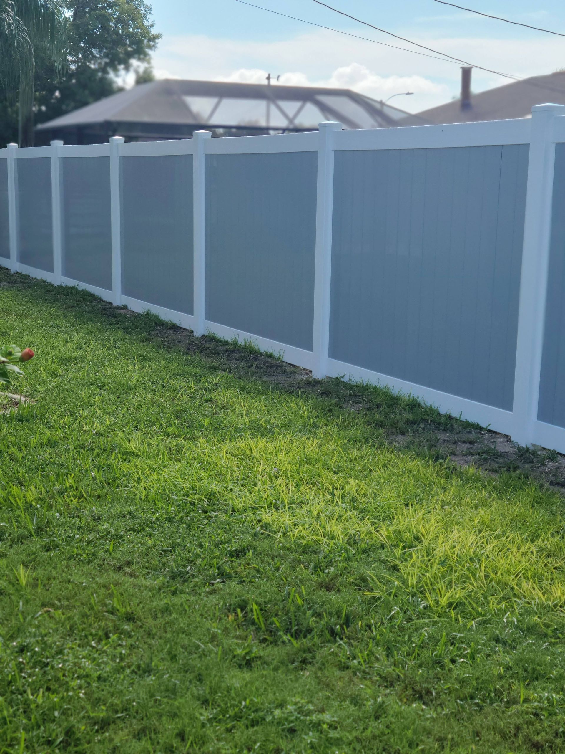 A gray and white vinyl fence borders a green grassy lawn under a sunny sky.