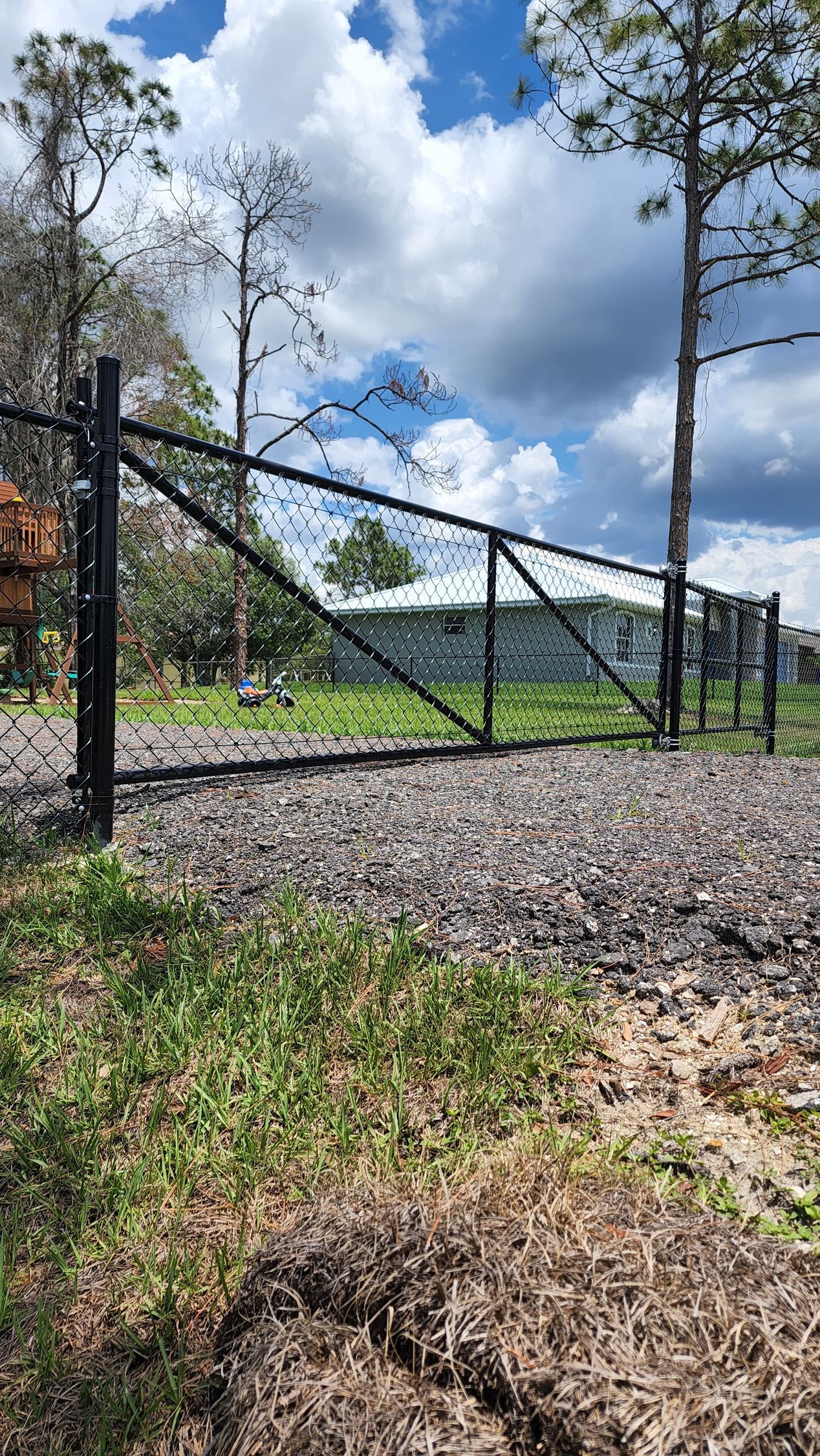 Black metal gate, open, across a gravel path. Green grass foreground, trees, blue sky with clouds.