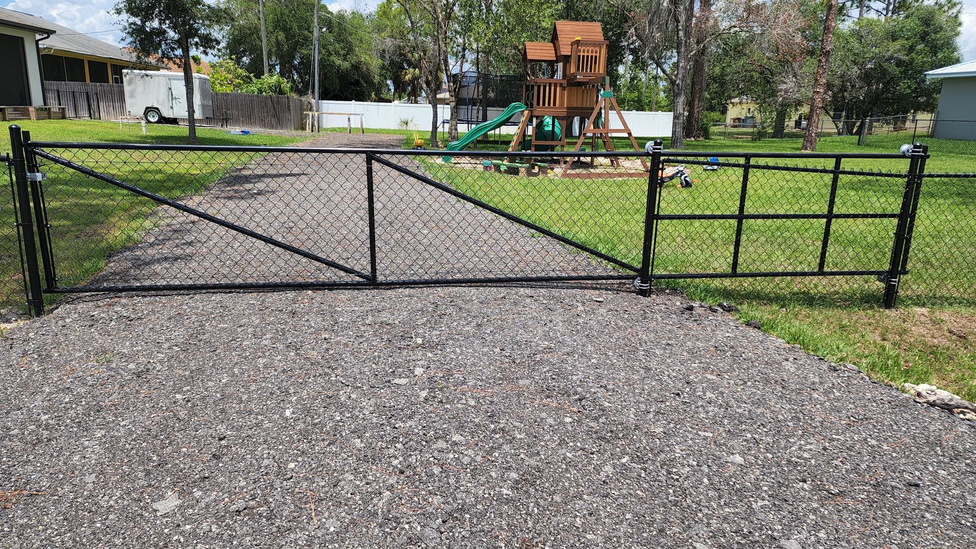 Black chain-link gate on a gravel driveway, leading to a yard with a wooden play structure.