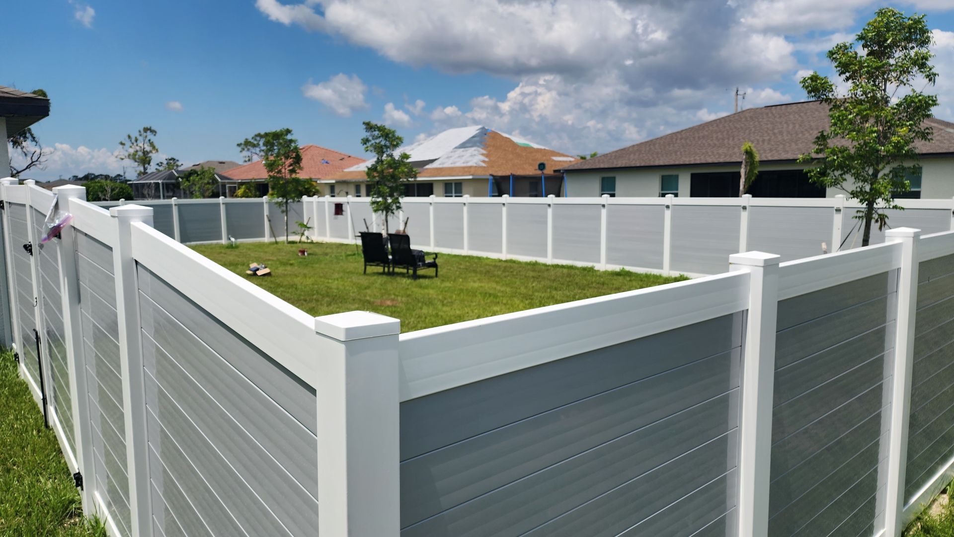 Fenced backyard with grass, two chairs, and surrounding houses on a sunny day.