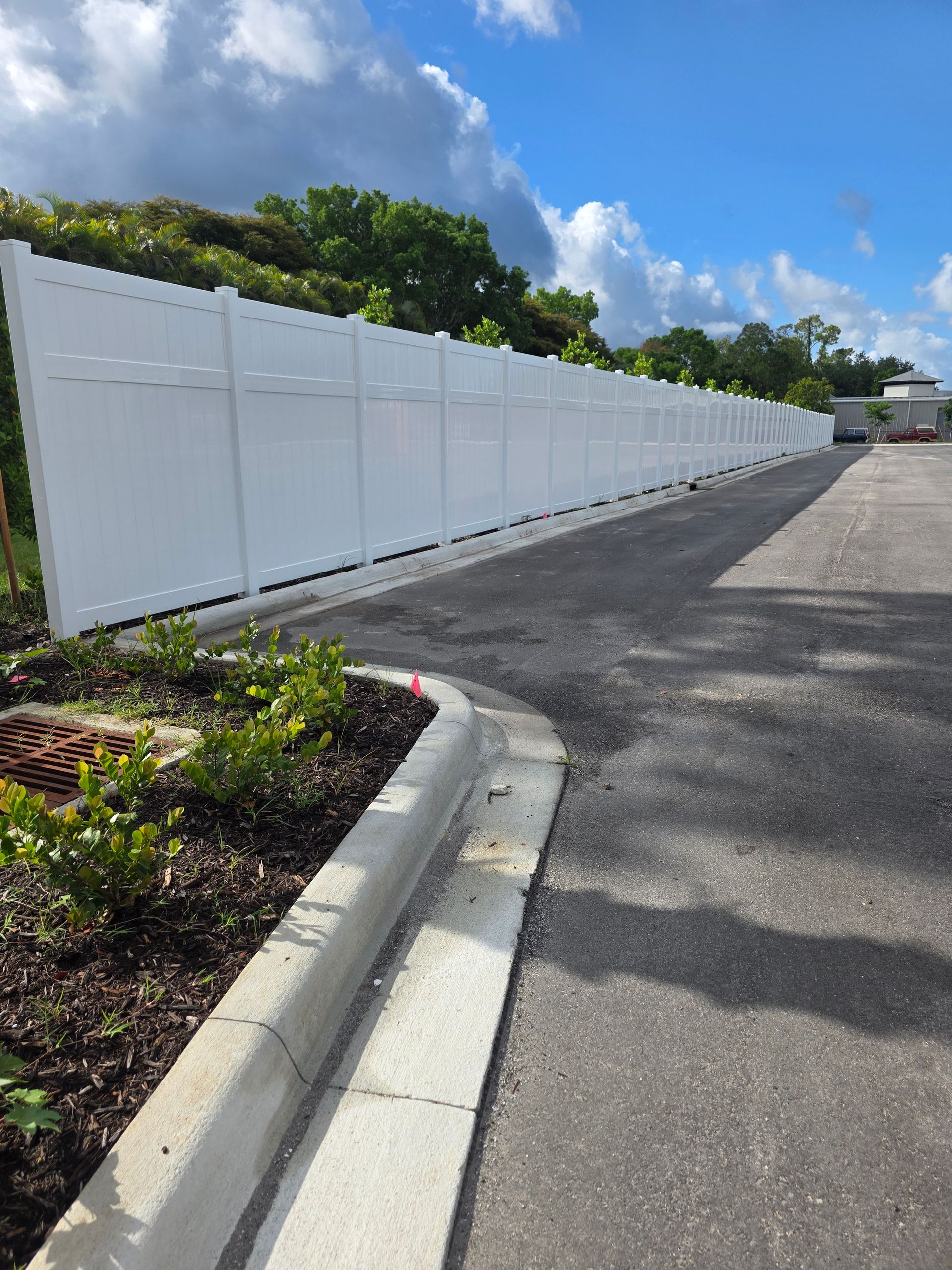 Long white fence along an asphalt road beside a curb and landscaped plants under a blue sky