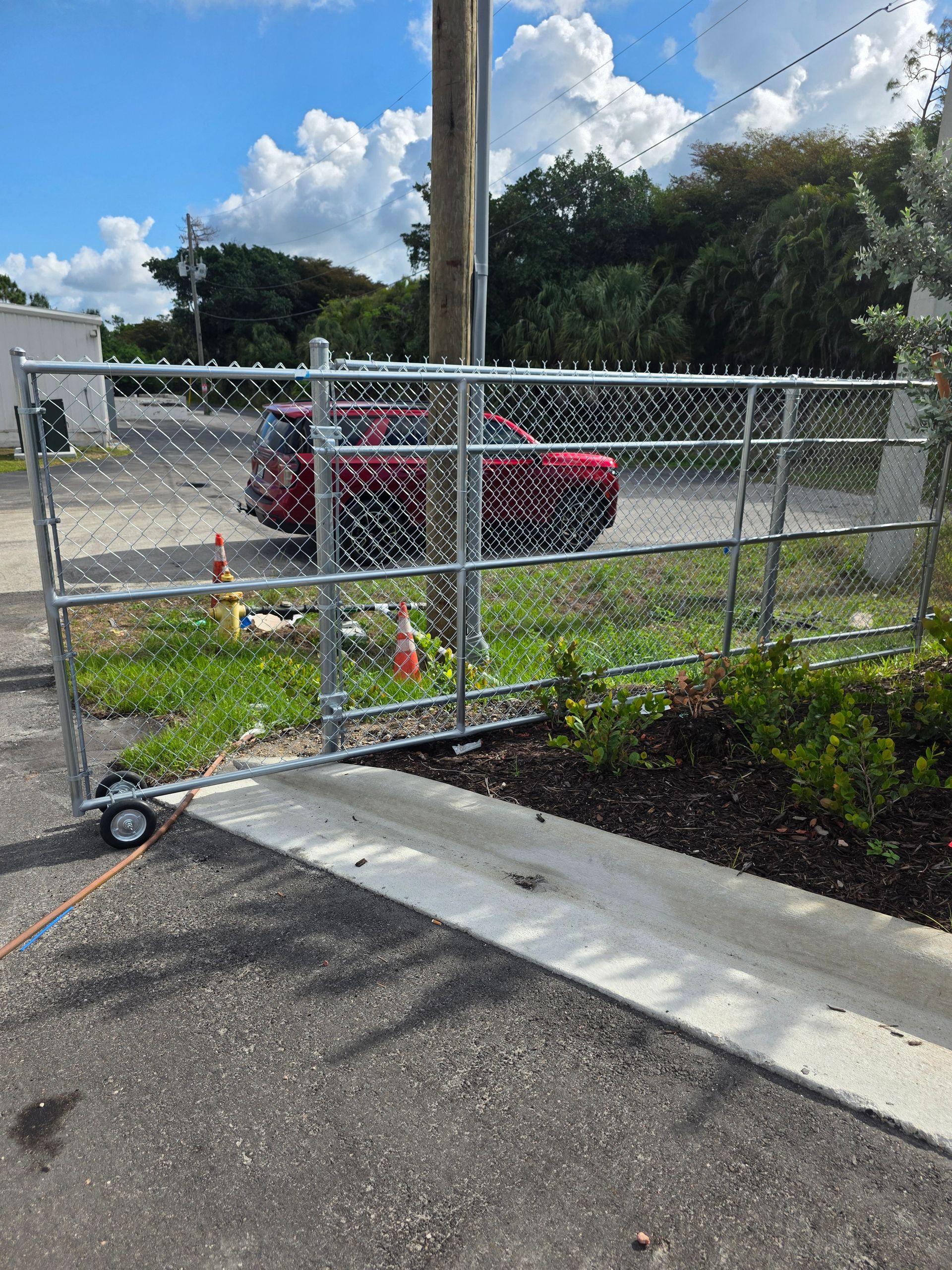Chain-link gate across driveway with red car behind it and trees under a cloudy sky
