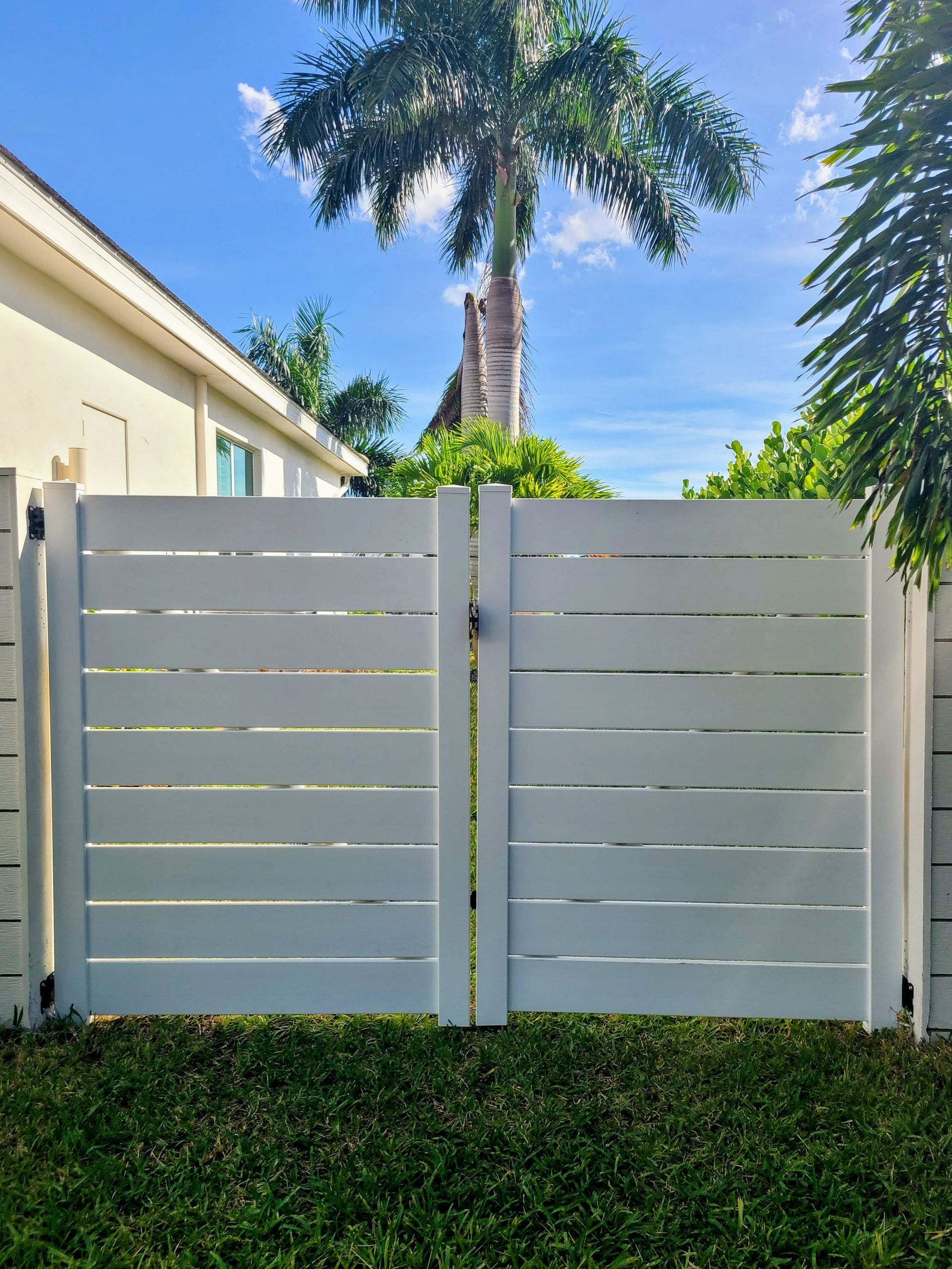White horizontal slat gate with a black lock, against a blue sky, flanked by a house and greenery.
