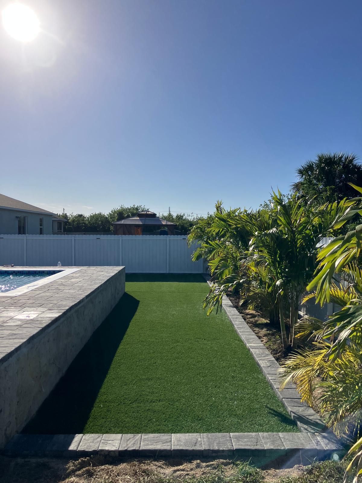 Sunny backyard with pool, artificial turf, and a white fence against a clear blue sky.