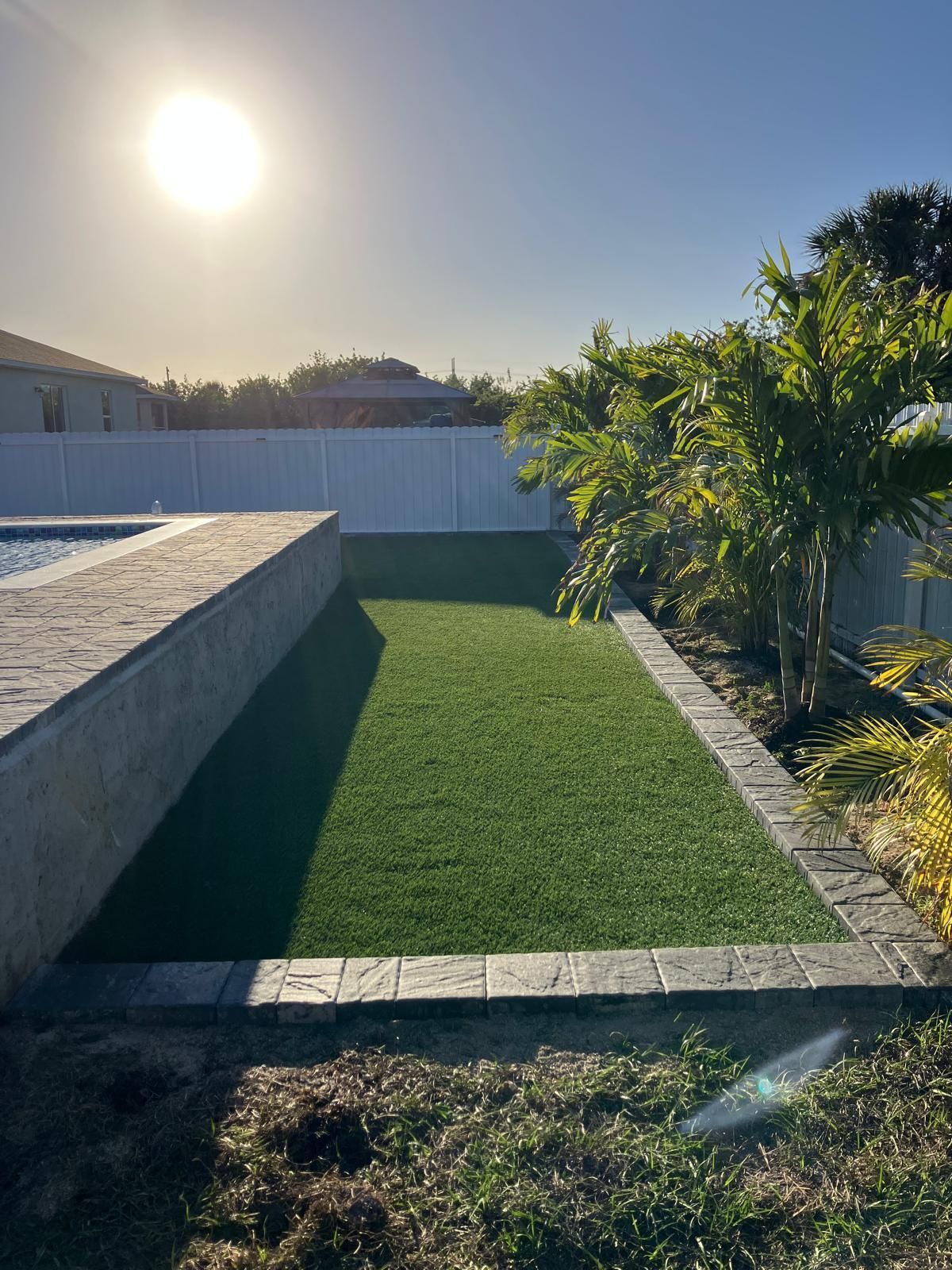Bright sun shines over a small lawn of artificial turf bordered by brick and a pool wall, with a white fence in the background.