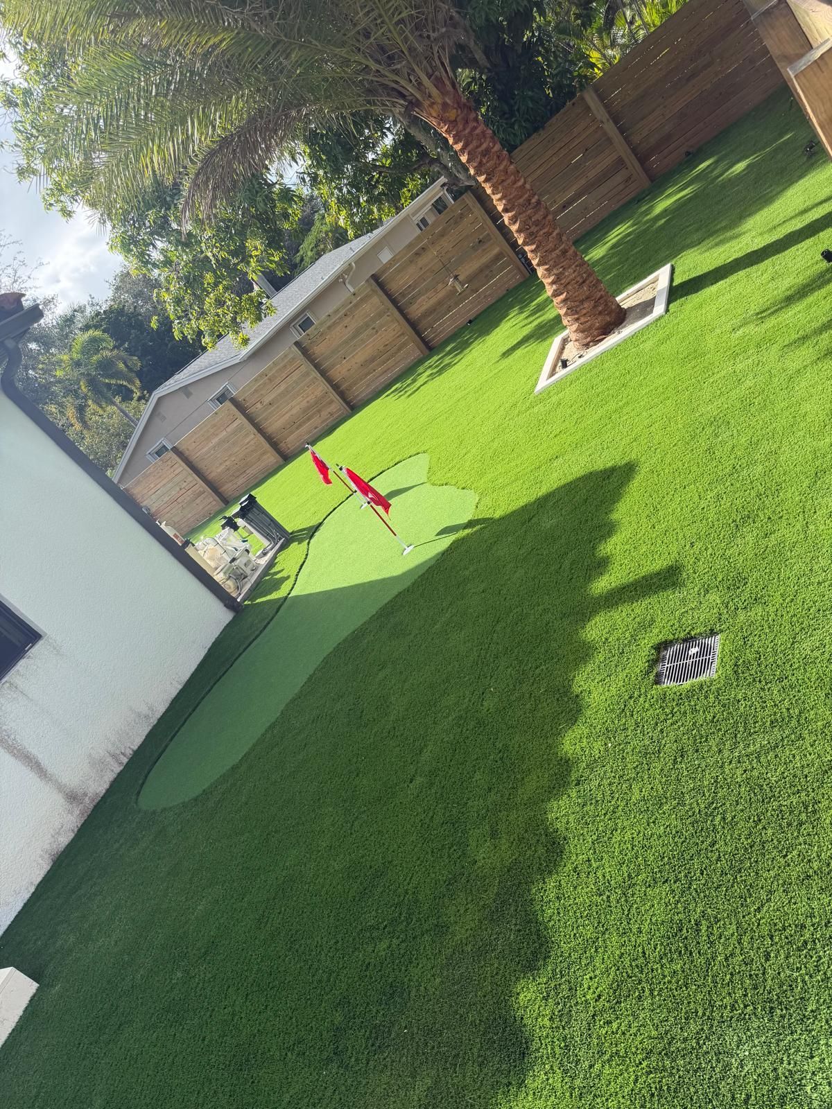 Backyard putting green with flags, surrounded by artificial grass, fence, and a palm tree.