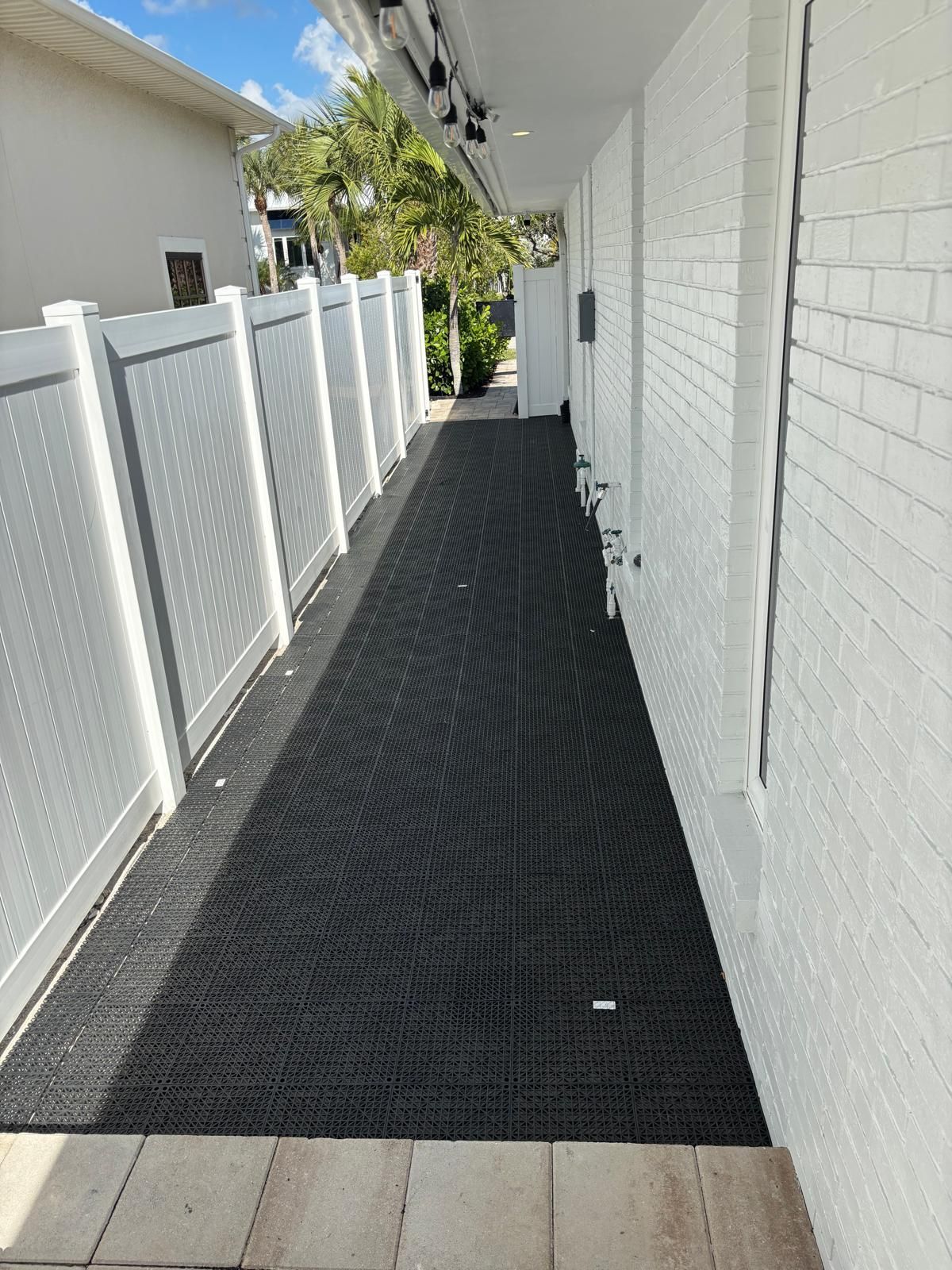 Narrow outdoor walkway with black grid flooring, white brick wall, and white fence.