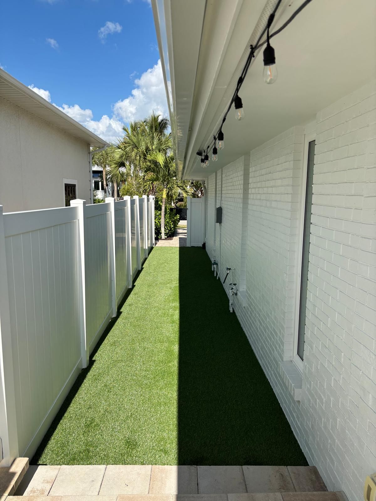 Narrow walkway with white walls and fence, green turf, string lights, and a sunny sky.