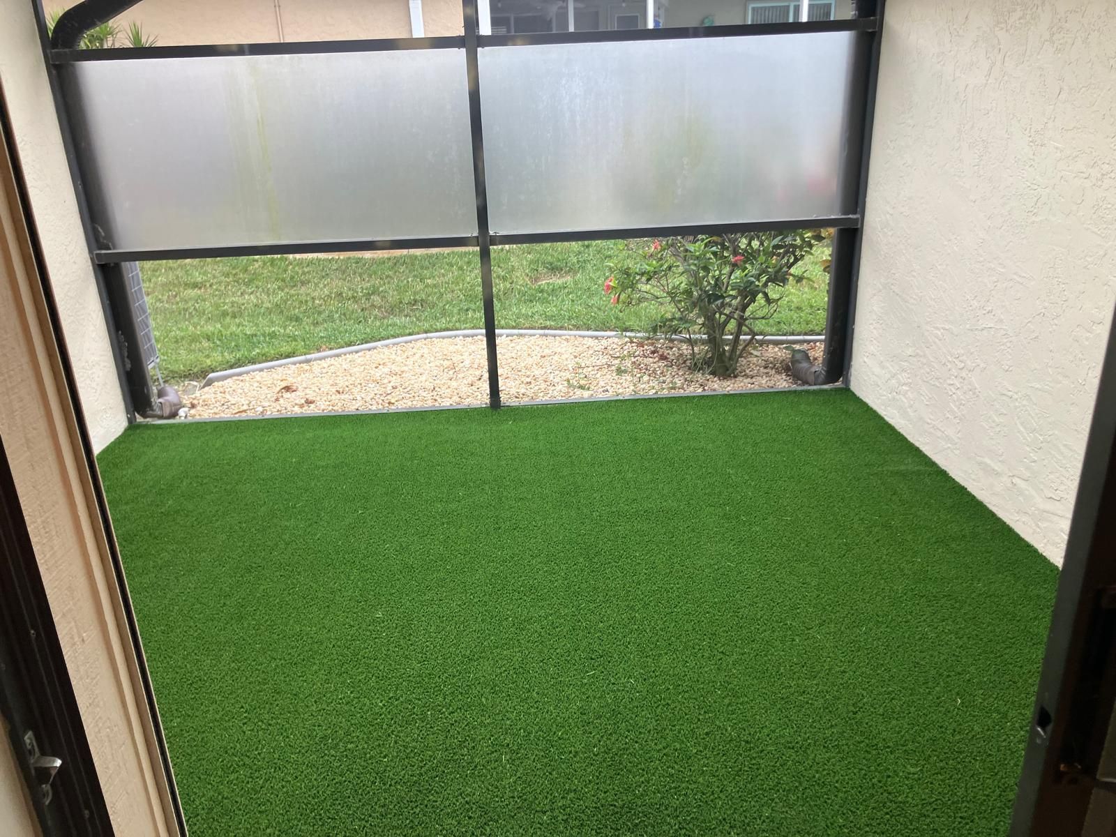 Small enclosed patio with artificial green turf and screened windows overlooking a grassy yard.