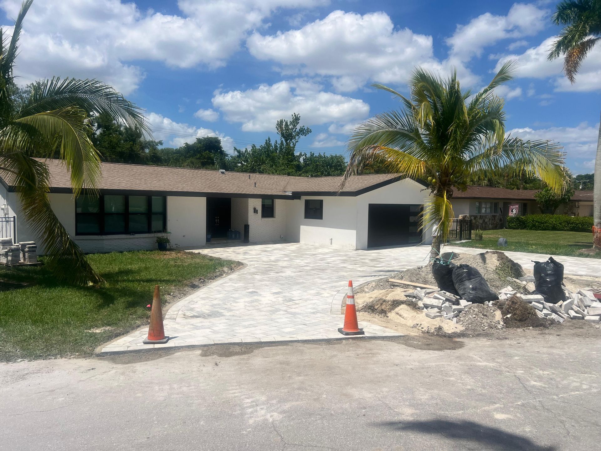 A single-story house with a white exterior, driveway construction, and palm trees under a blue sky.