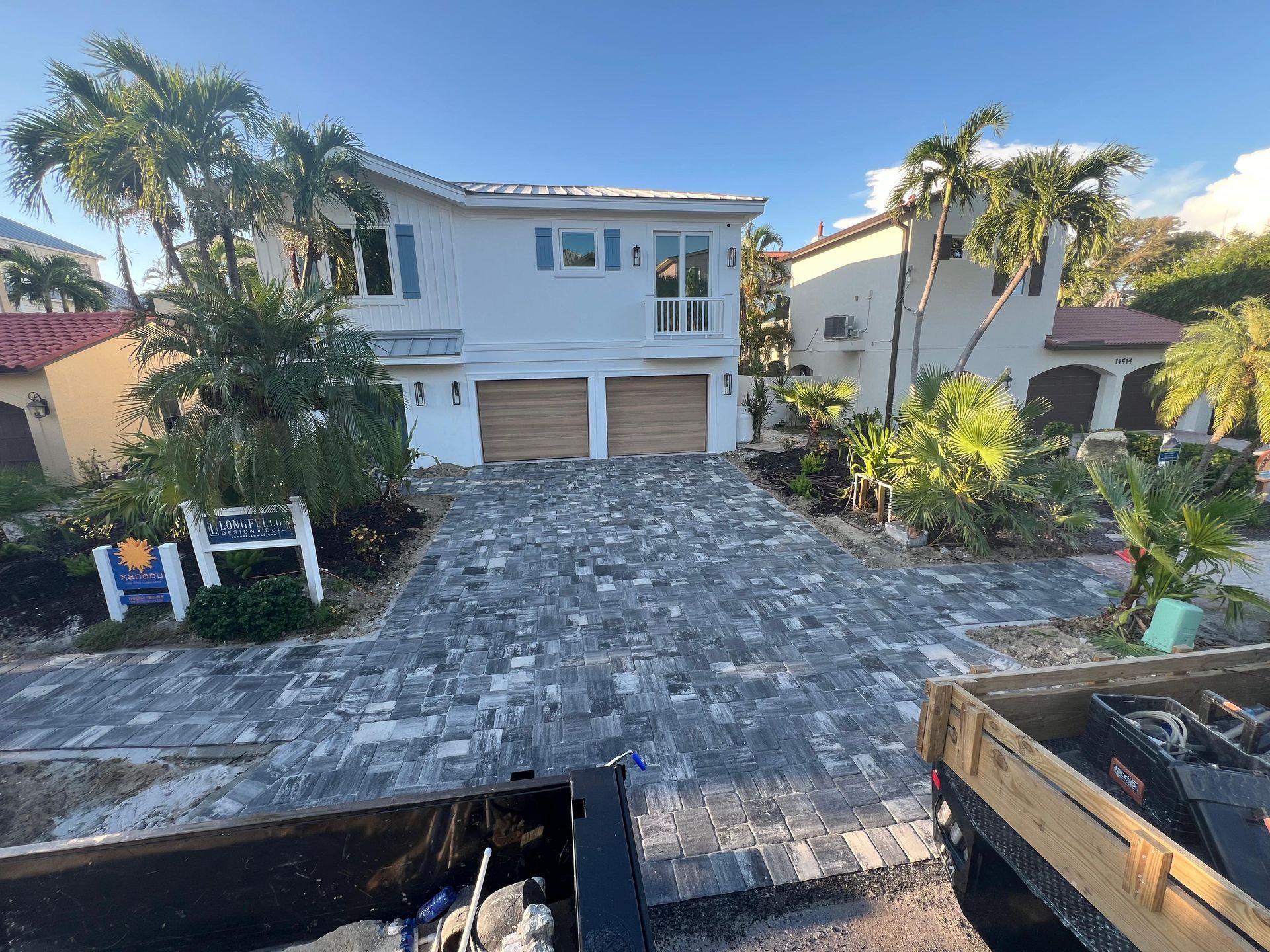 House with grey paver driveway, two-car garage, and palm trees under a blue sky.