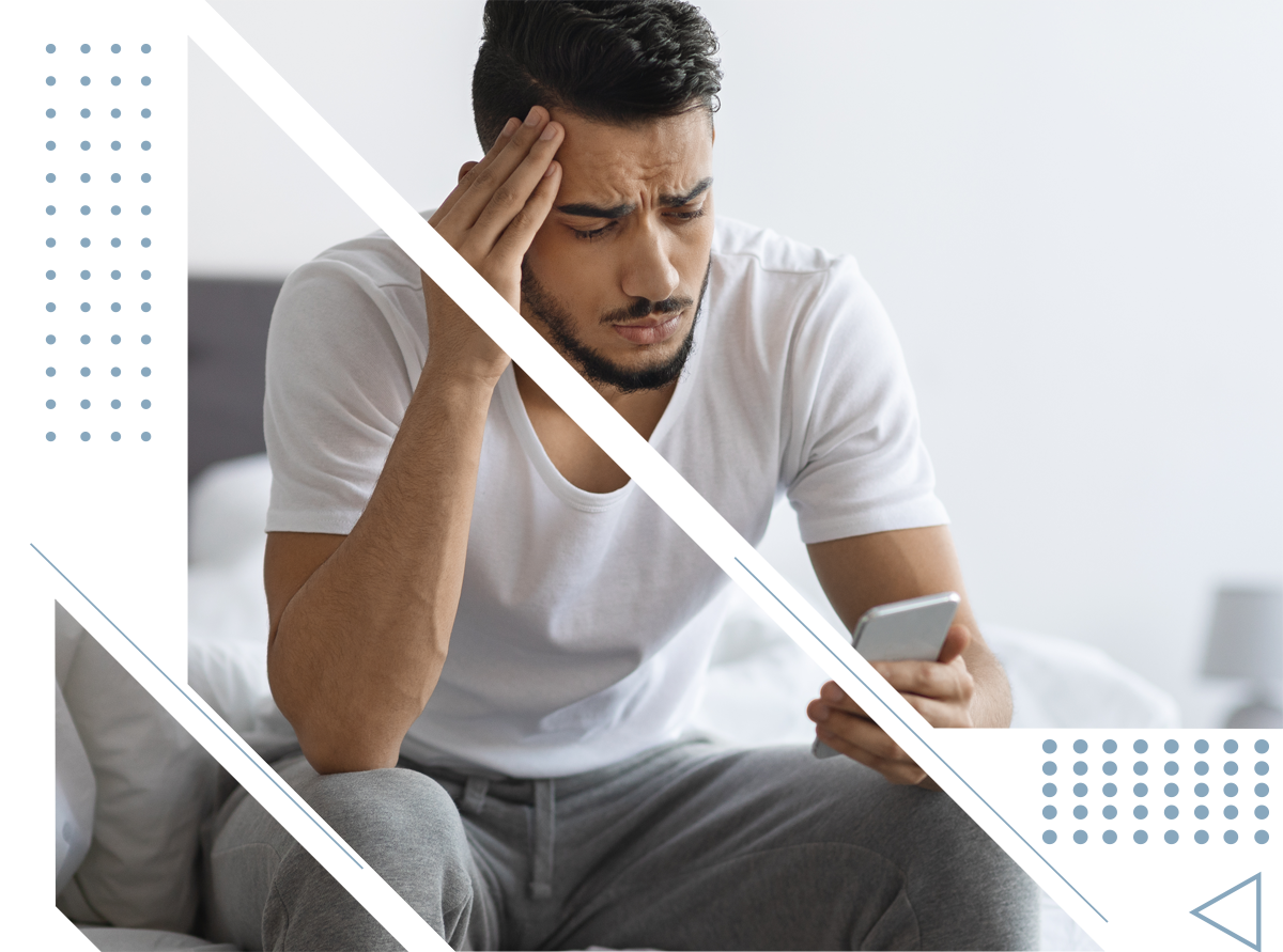 A man is sitting on a bed holding his head while looking at his phone.