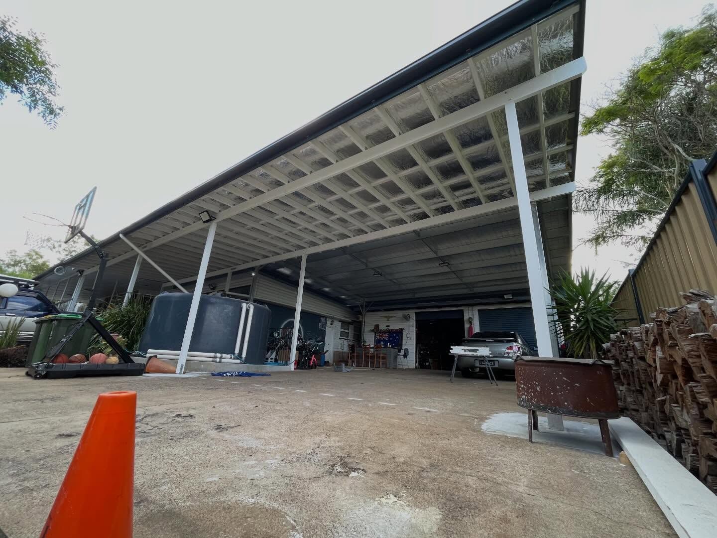 A wide-angle view of a large, open-air carport featuring a metal roof, supporting white beams, and a concrete driveway.