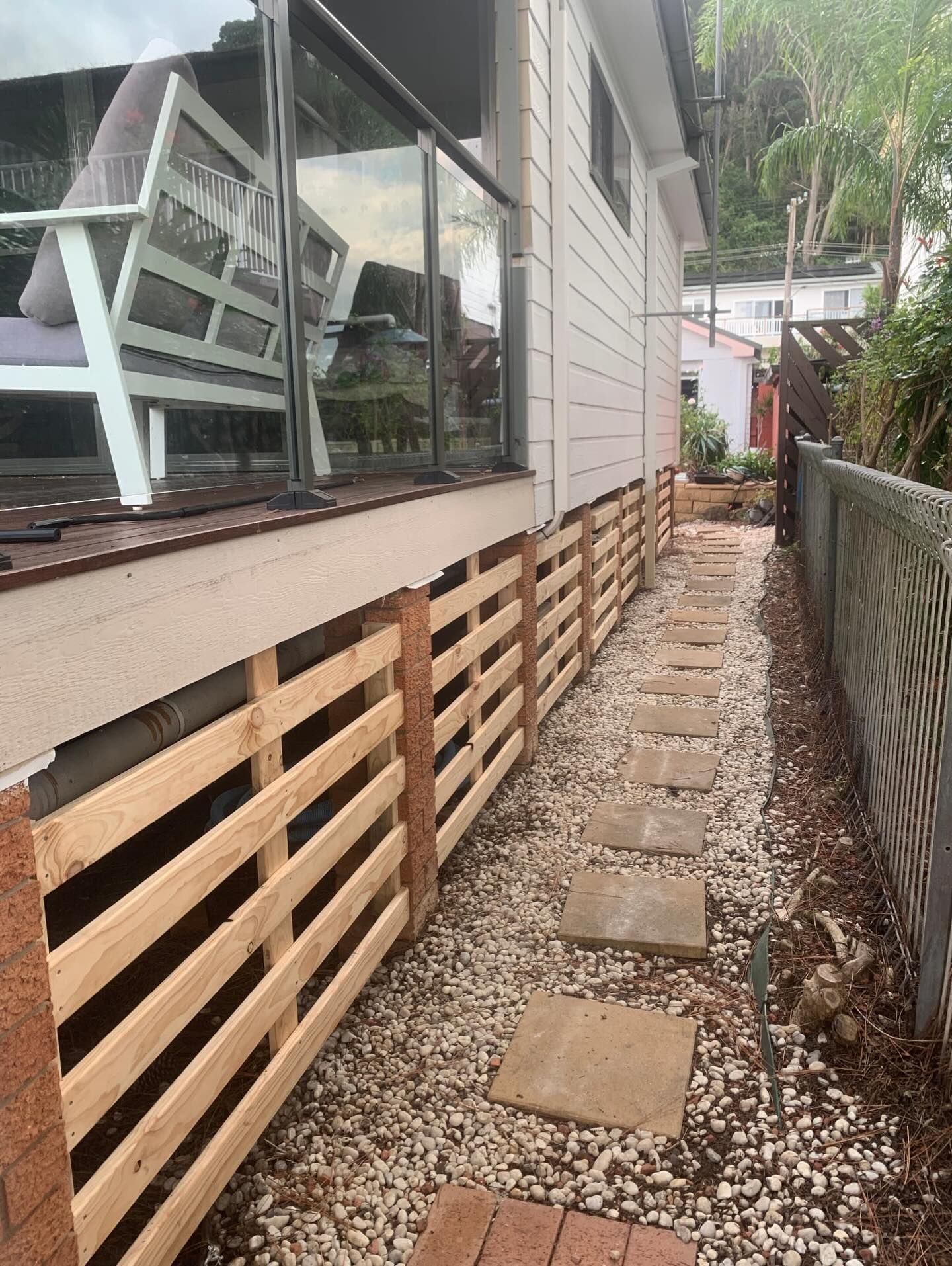 A side view of a house with a wooden deck featuring a slatted privacy screen above a gravel path with stone stepping tiles.