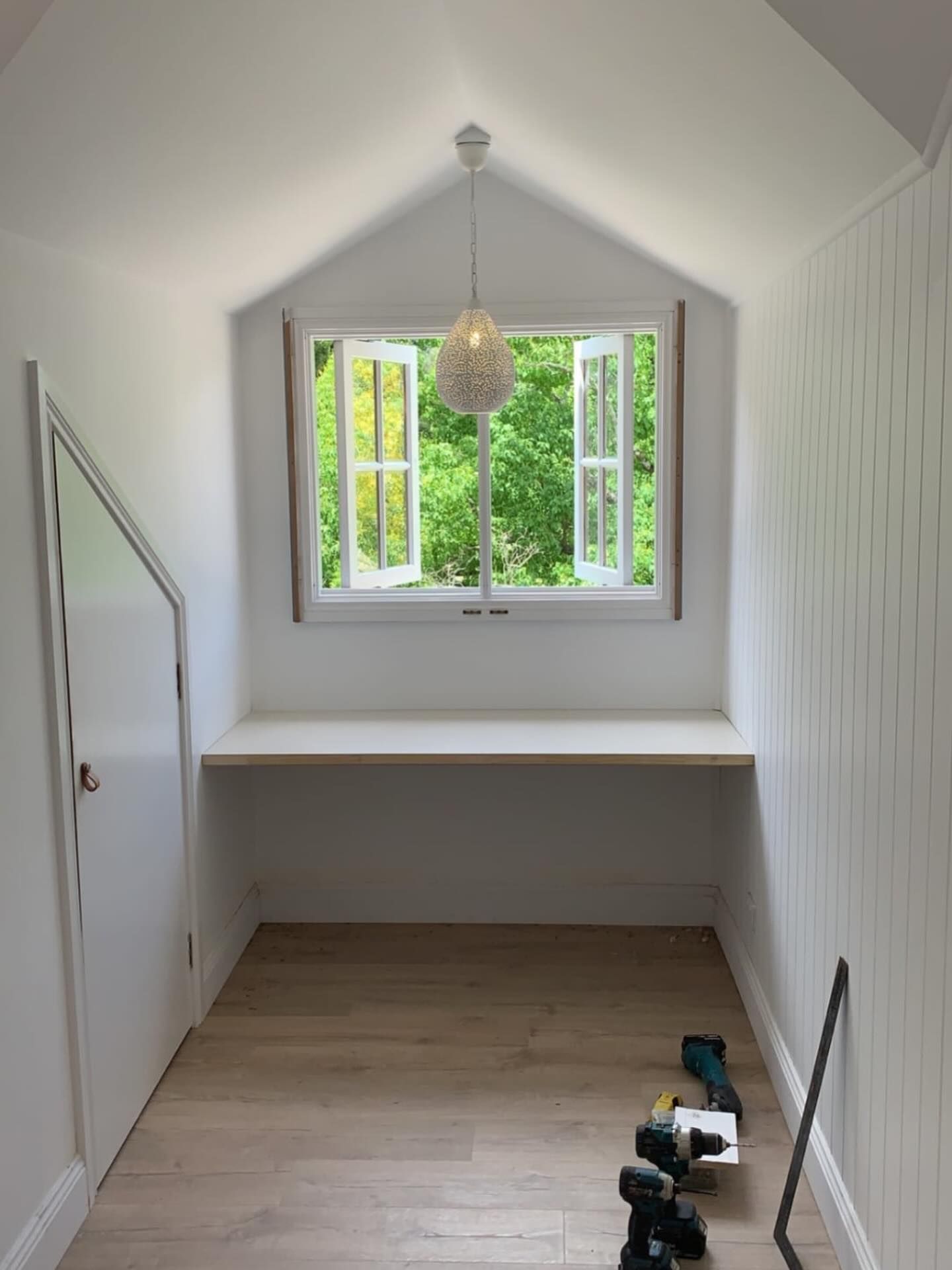 A small, white, sunlit office nook with a built-in desk under a window overlooking green trees, with tools on the floor.