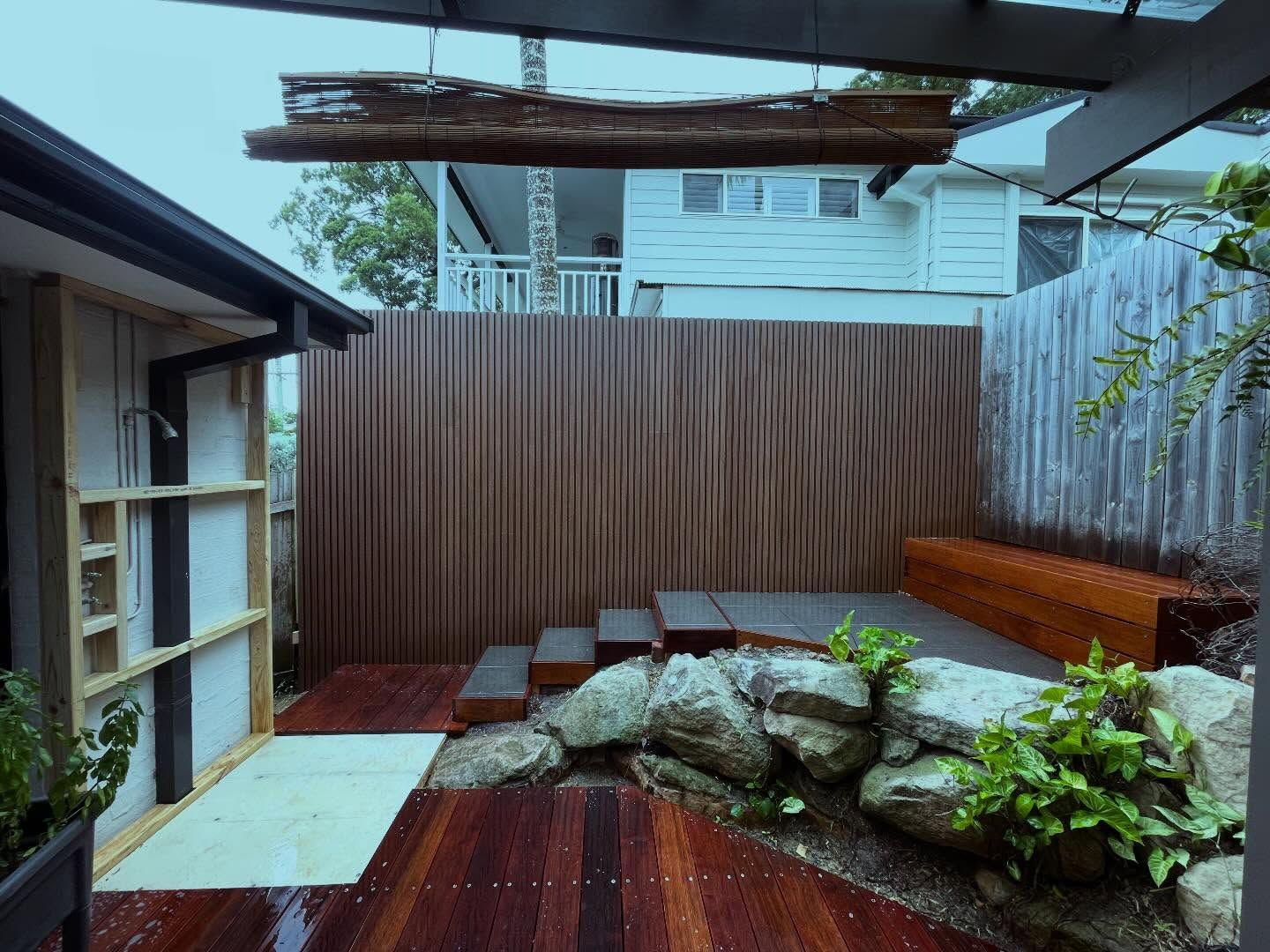 An outdoor patio with wooden decking, stone landscaping, a built-in bench, a wooden slat privacy wall, and an outdoor shower.