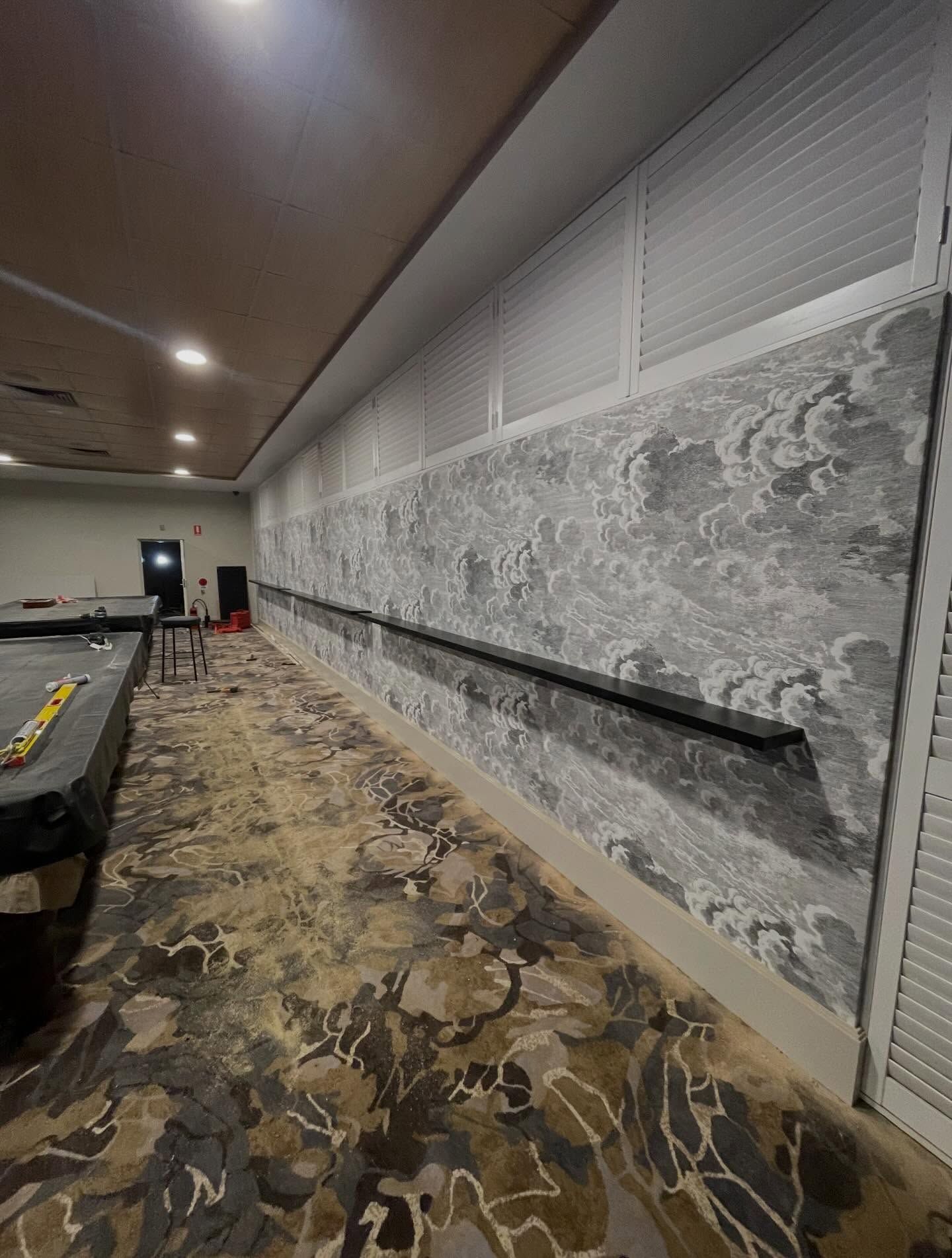 A long, grey patterned wall features a thin, dark horizontal shelf. A work area with a yellow level sits on the left.